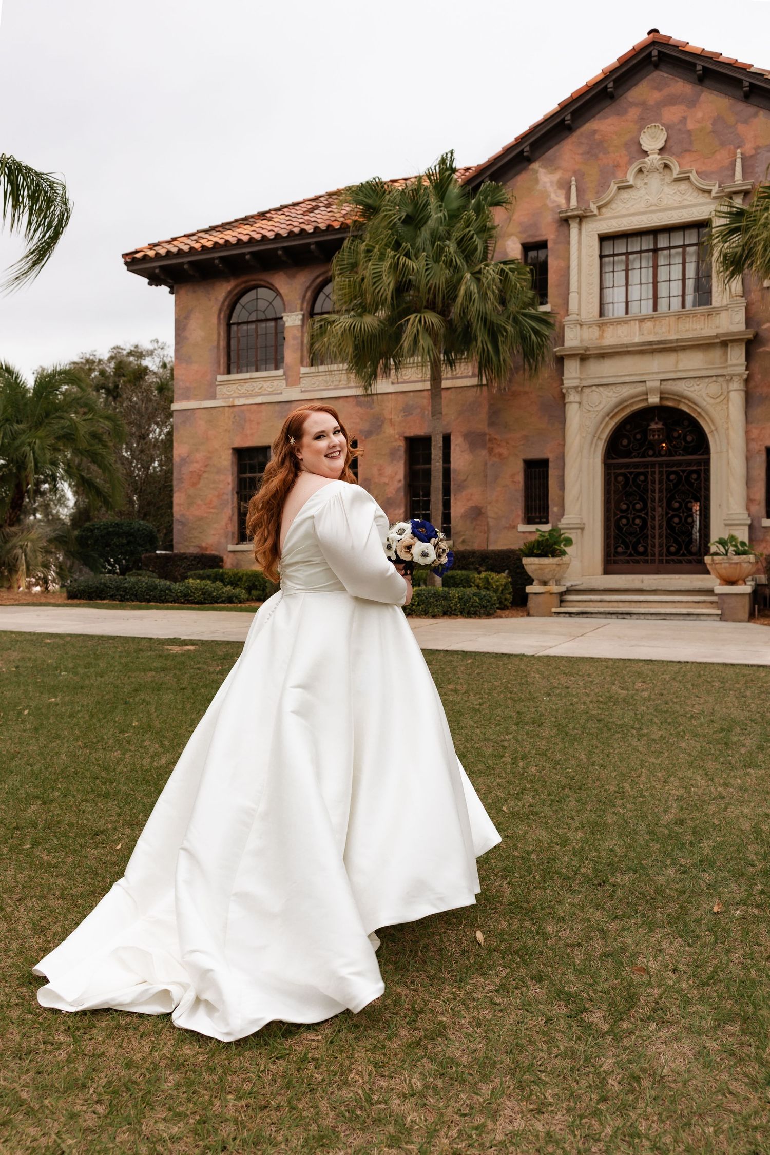 Smiling redhead bride in white satin gown holds bouquet outside elegant Spanish estate.
