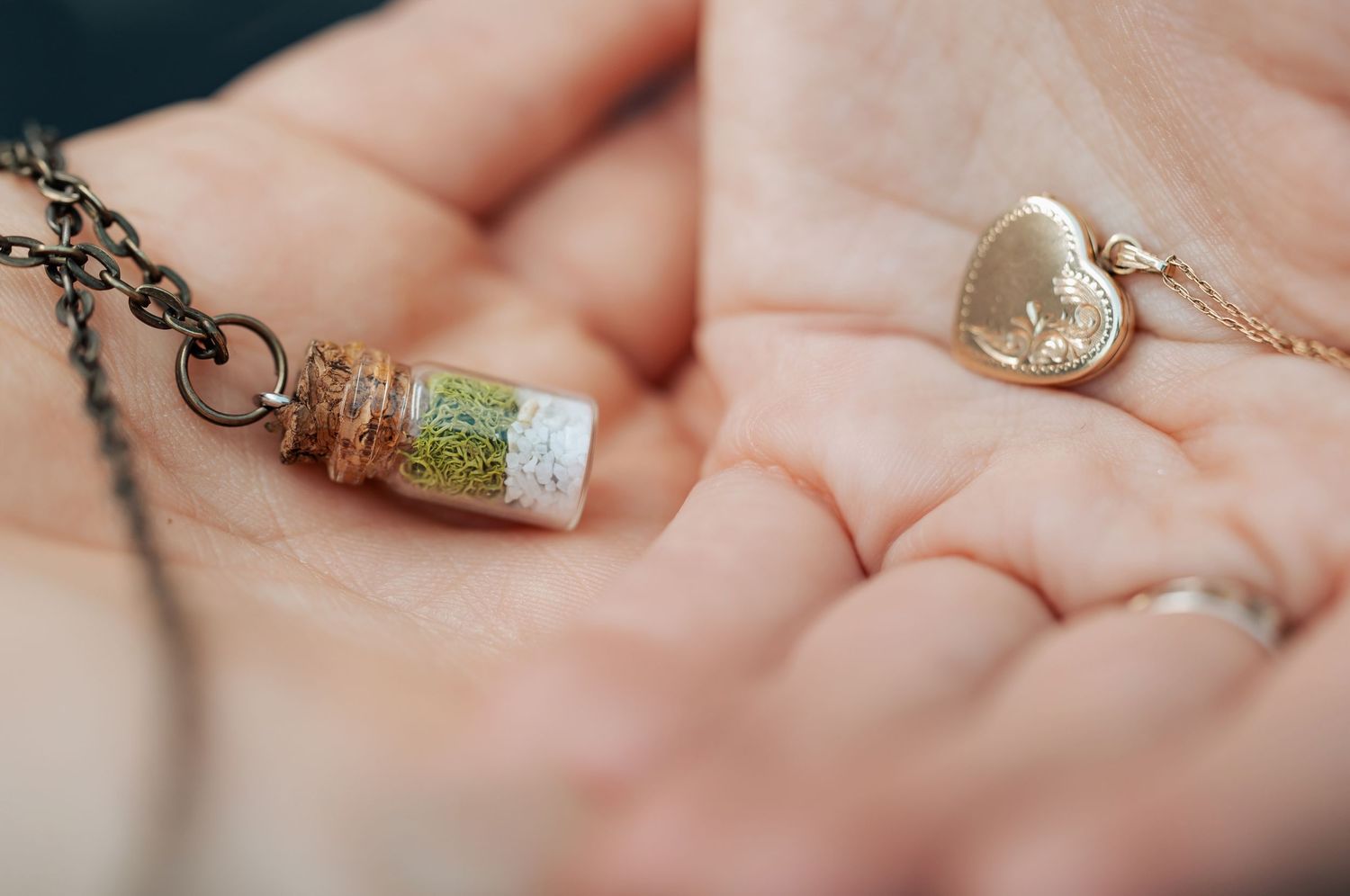 Two hands hold delicate necklaces, one a tiny glass bottle pendant and one a gold heart locket.