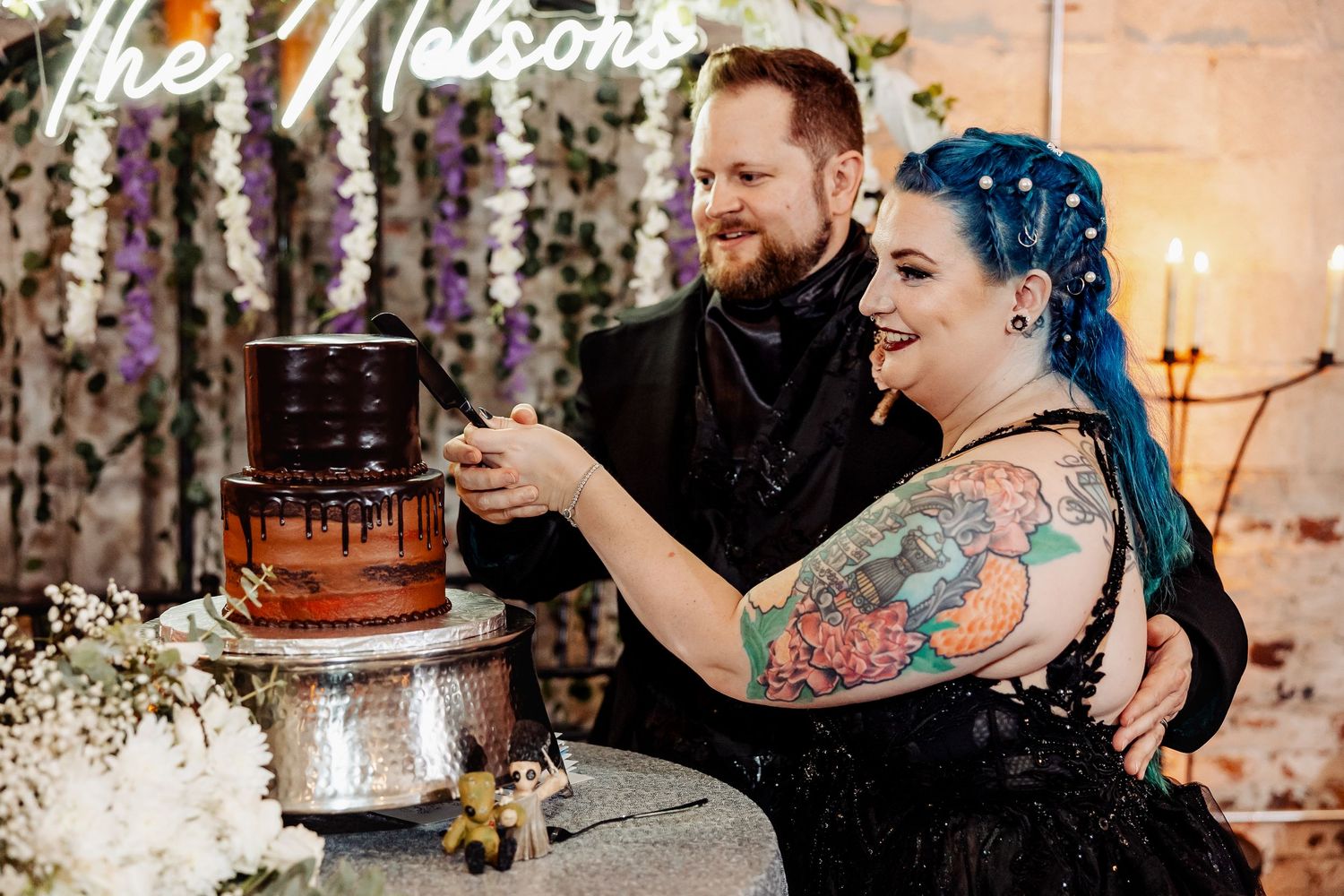 Couple cutting a dark chocolate drip wedding cake beneath a neon Nelsons sign at their reception.