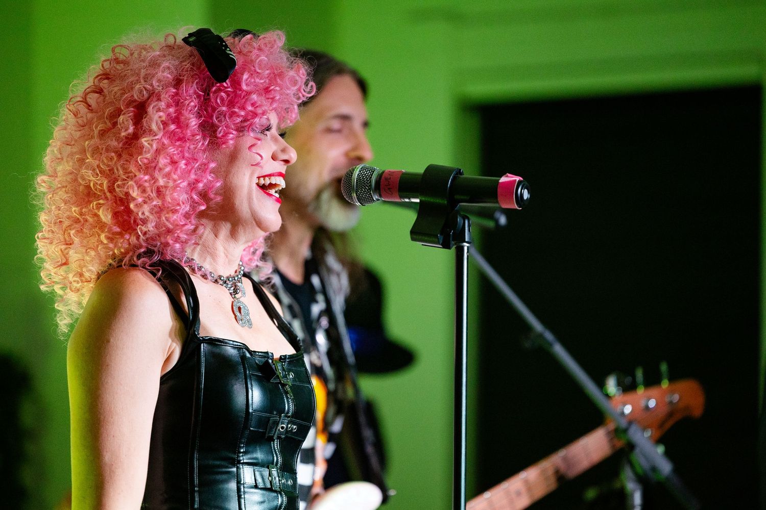 Woman with pink curly wig singing at microphone with guitarist performing on stage at live music event.