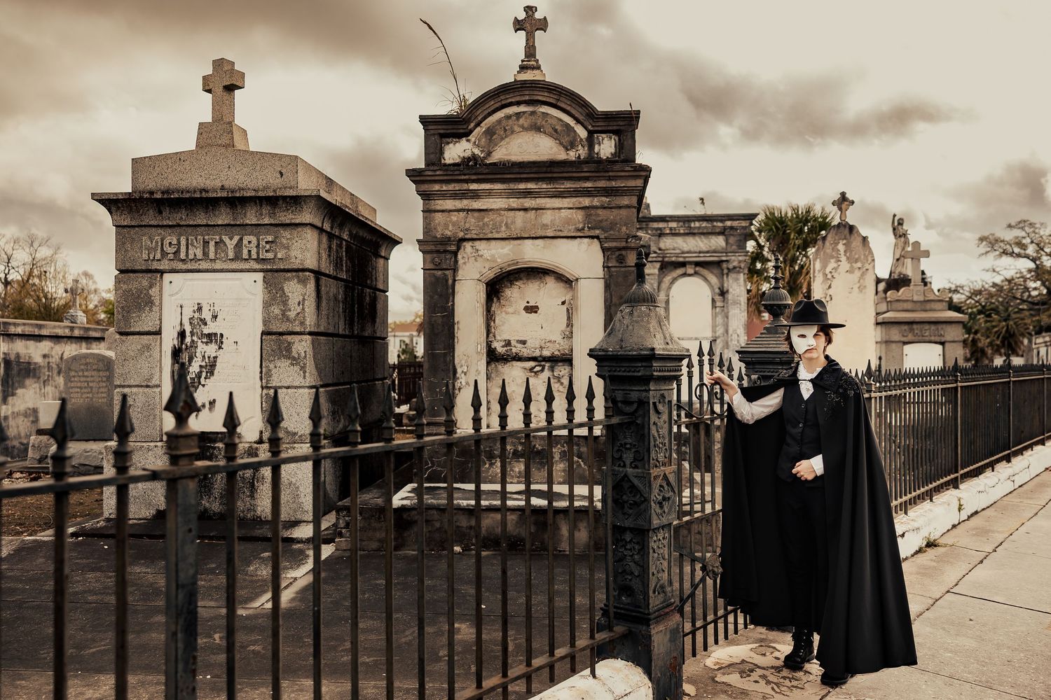 Gothic figure in black cape and hat standing beside ornate cemetery tombs in New Orleans.