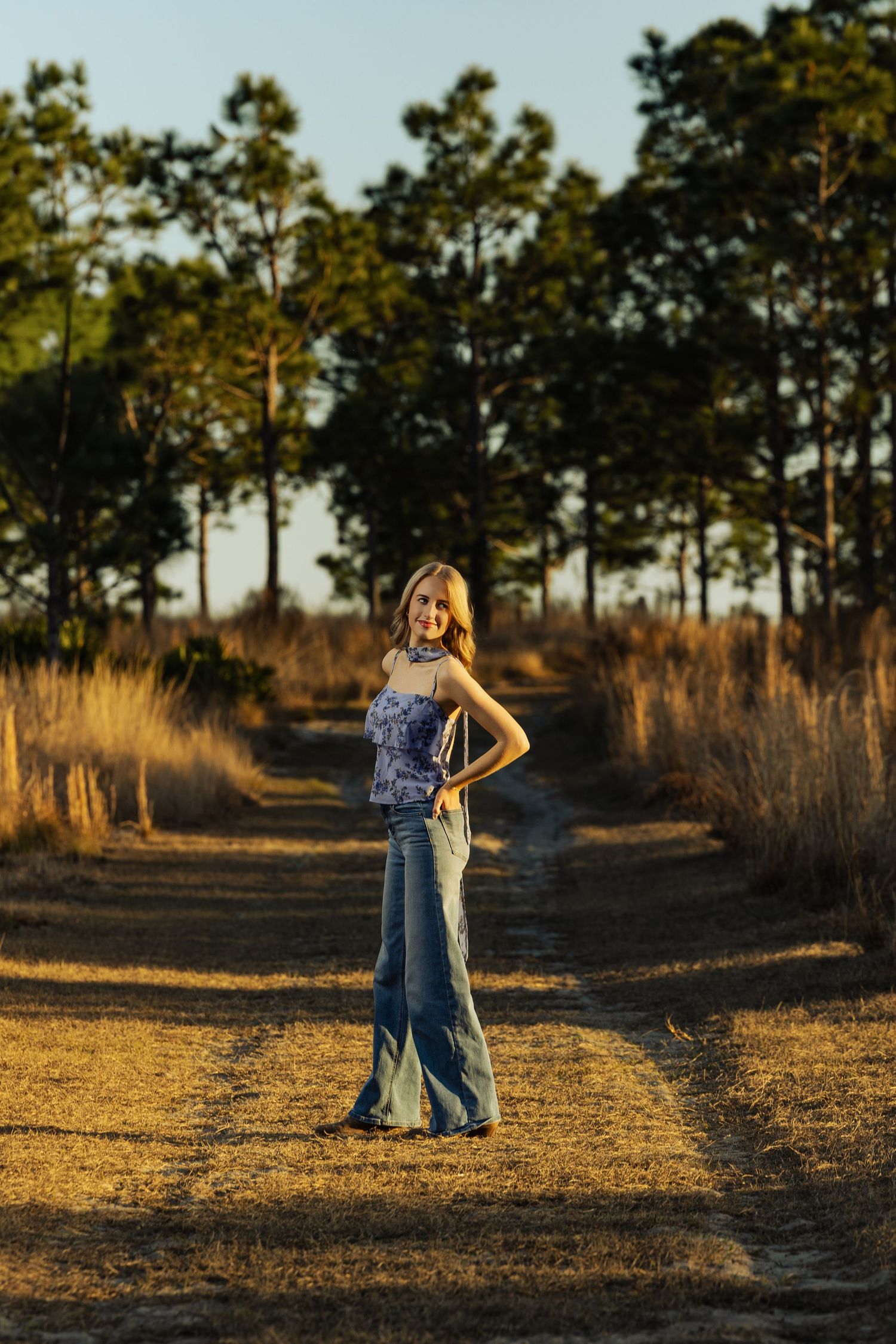 Young blonde woman in flared jeans posing on a rural dirt path surrounded by golden grasses at sunset.