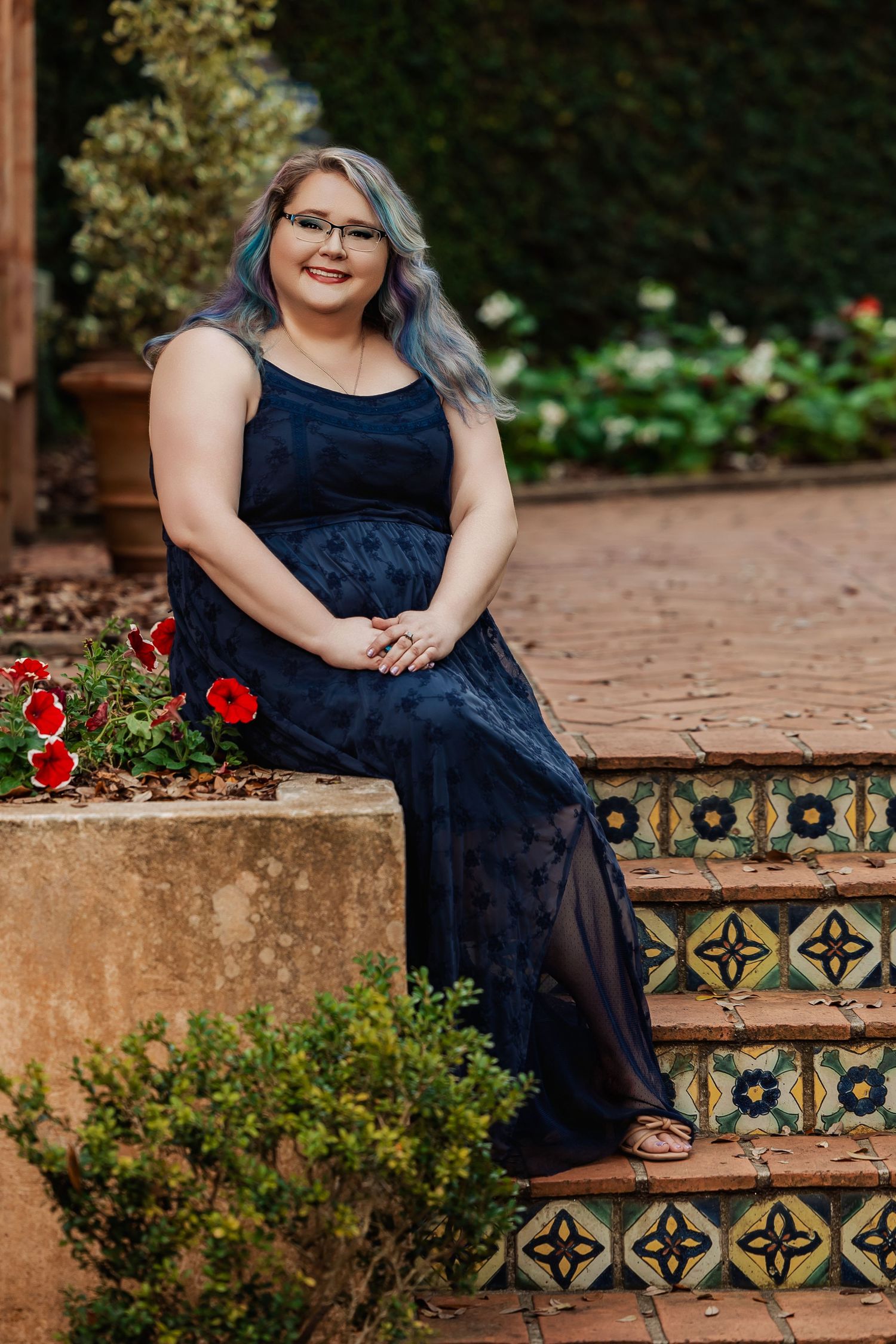 A young woman with blue-gray hair in a navy lace gown sits on decorative tiled steps in a garden.