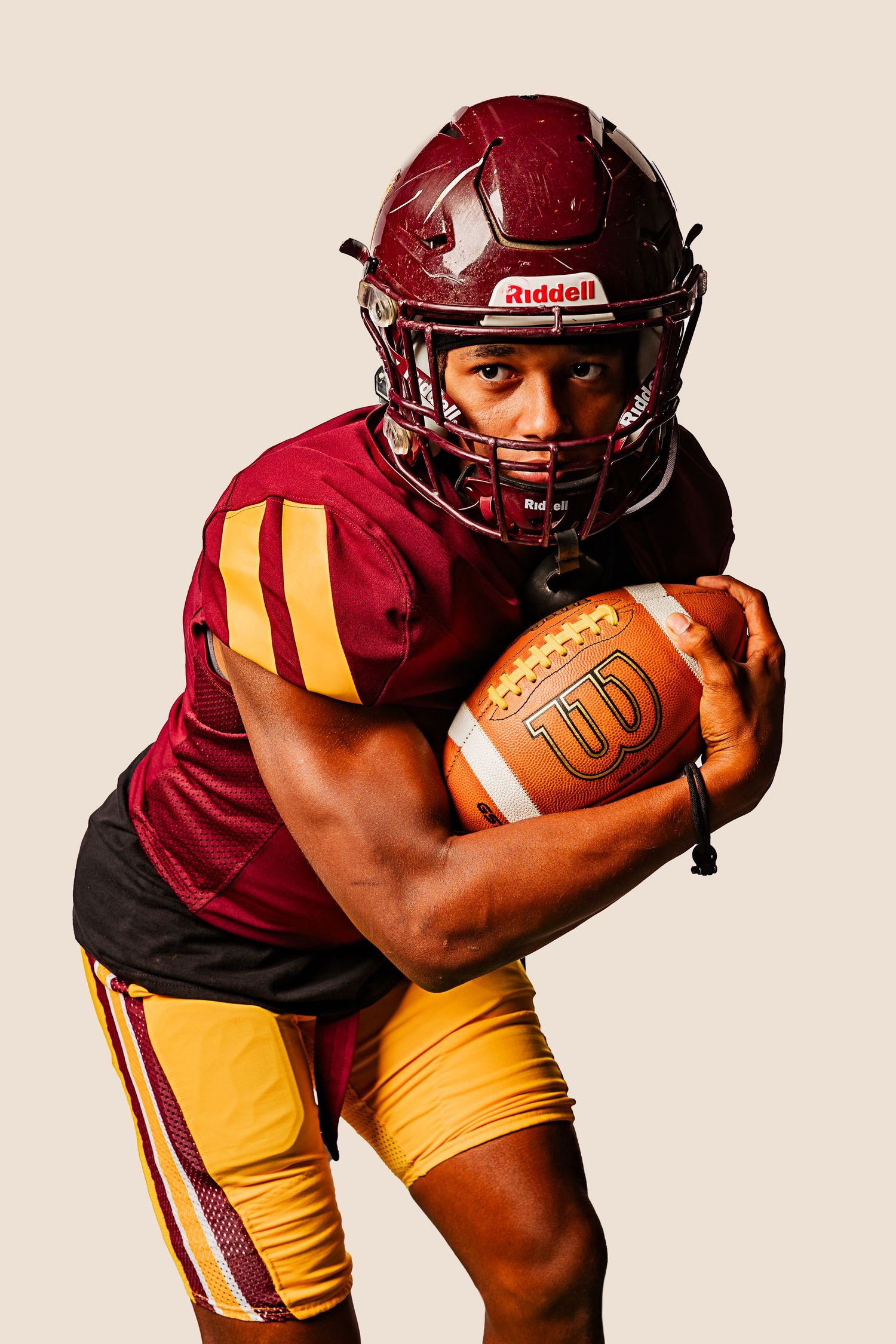 Football player in maroon and gold uniform holding a Wilson football, wearing a Riddell helmet, in a dynamic pose.