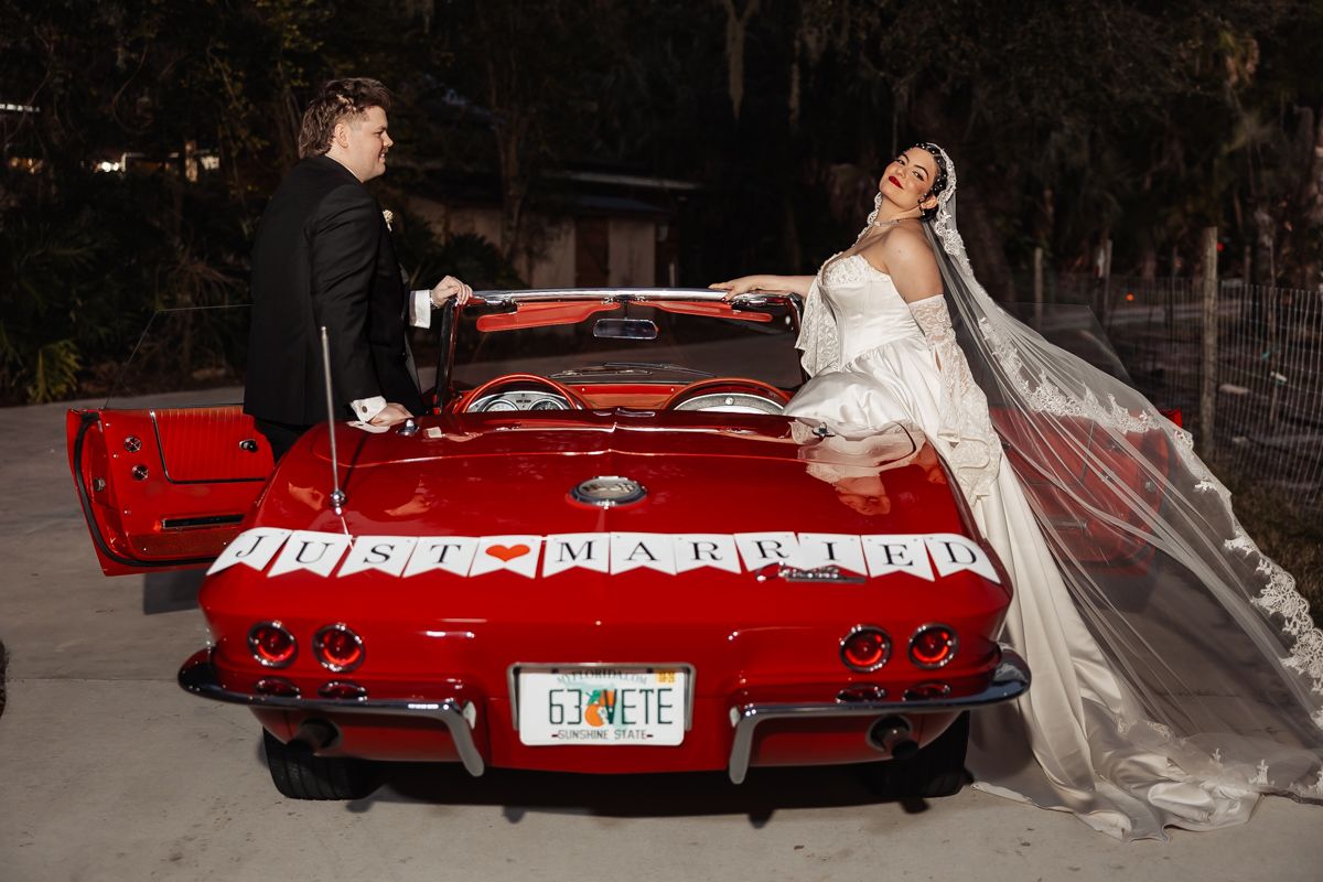 Bride and groom posing with a red vintage Corvette decorated with Just Married sign at night.