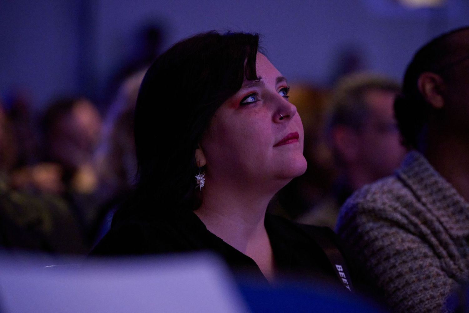 A person watches attentively in a dimly lit audience with blue lighting in the background.