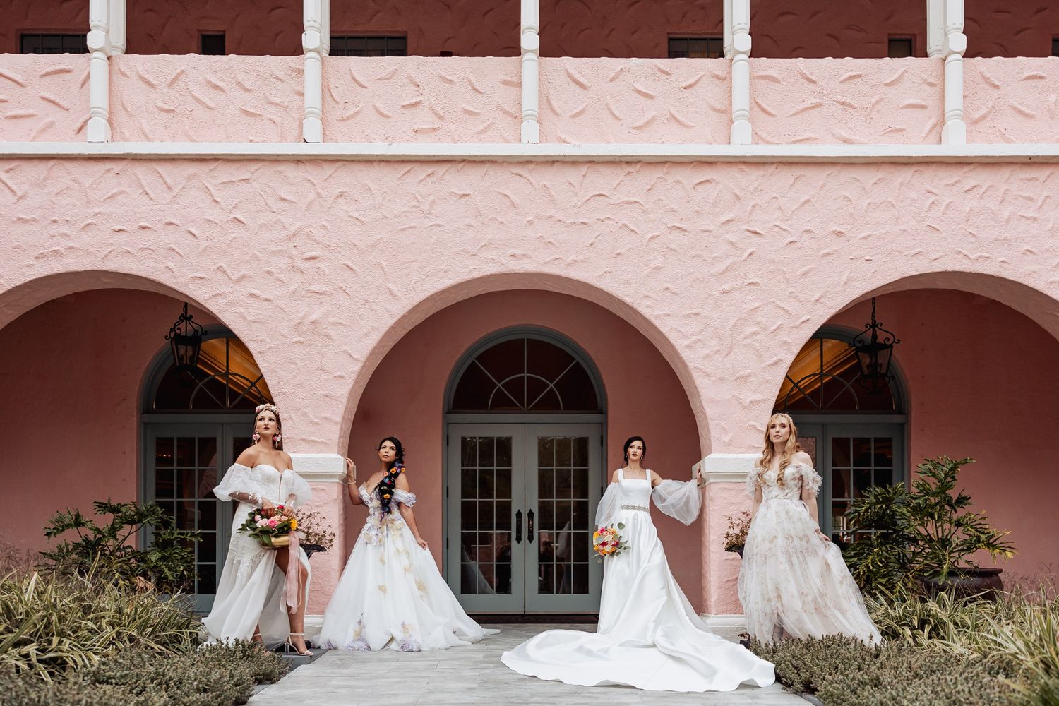 Four people in white formal dresses stand in front of a pink Spanish-style building with arched windows and doorways.