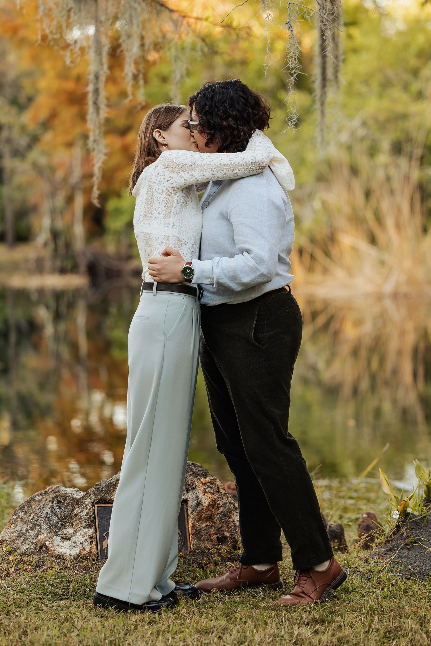 Two women share a romantic kiss while embracing near a serene autumn pond with colorful foliage.