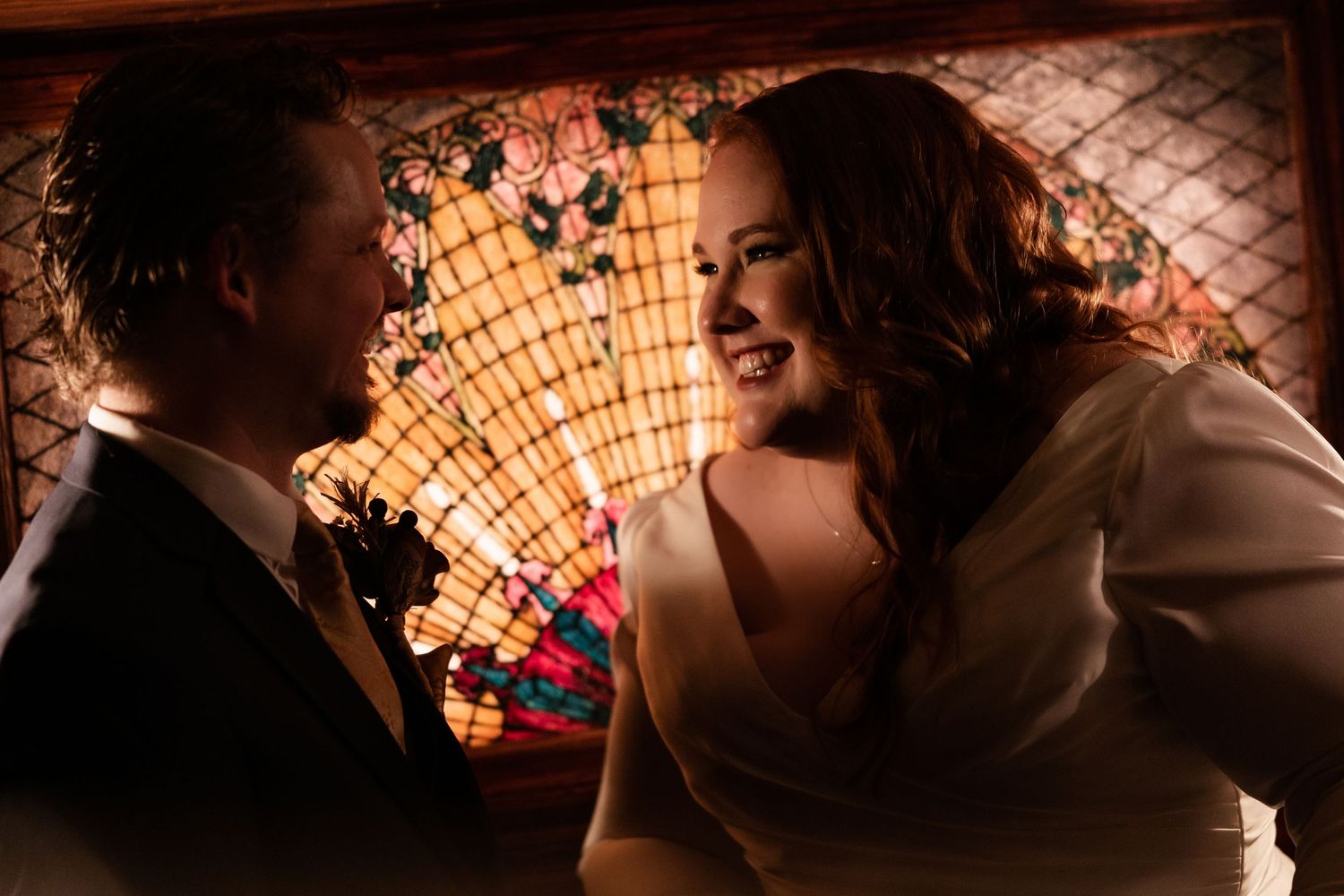 Bride in white gown smiles at groom beside colorful stained glass window in warm light.