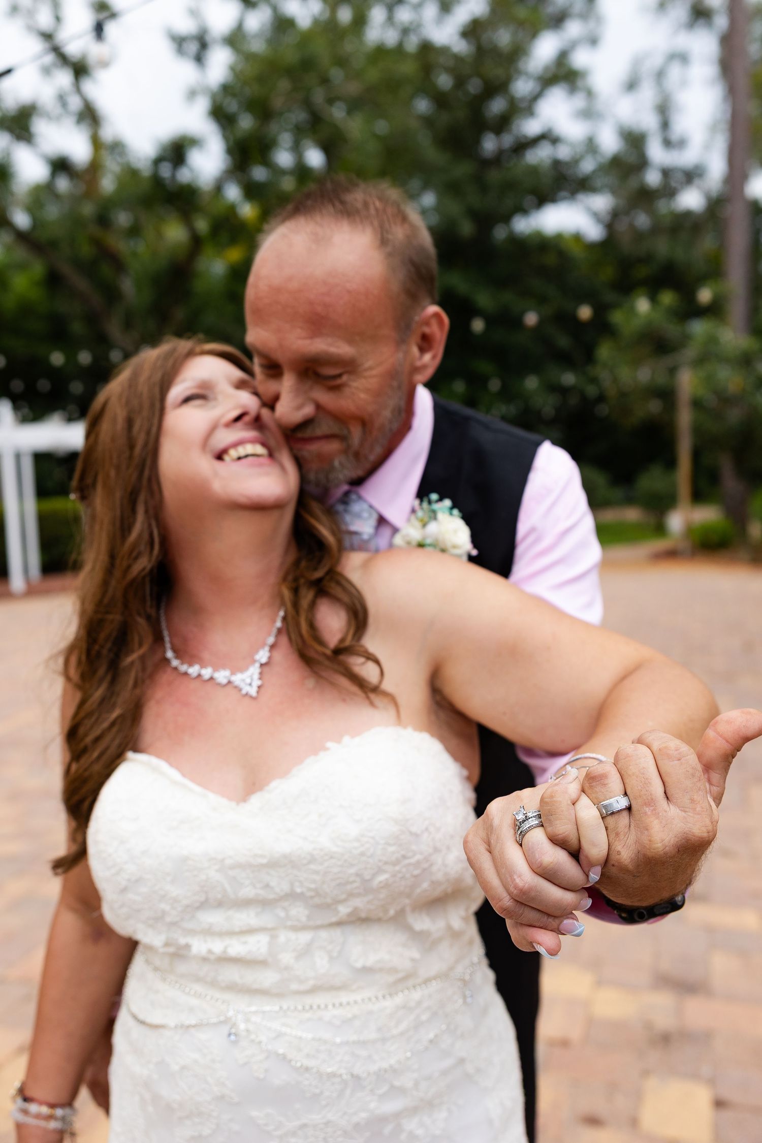 Groom kisses bride on cheek while showing off wedding rings, both smiling joyfully on brick patio.