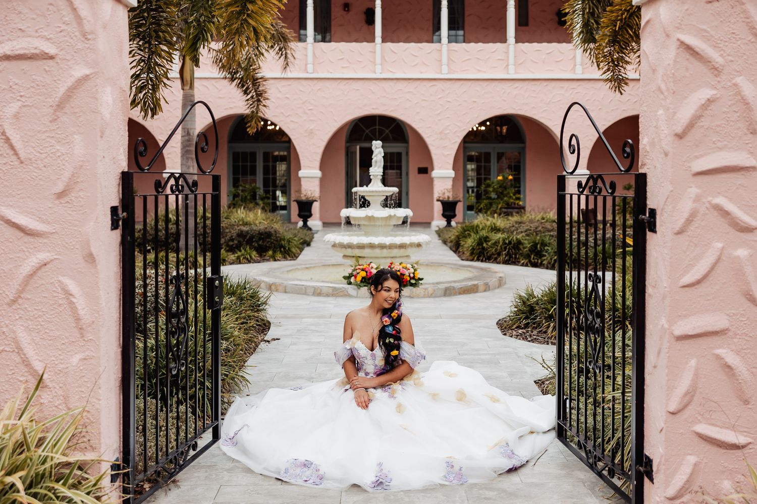 Bride in white ball gown sits gracefully at open iron gates of pink Spanish-style courtyard venue.
