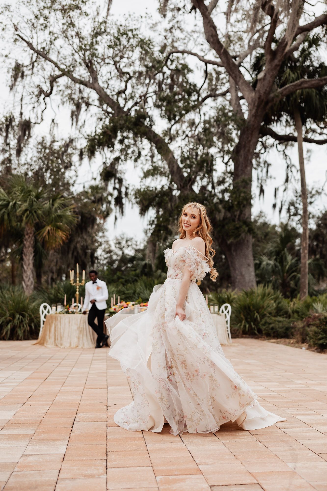 Blonde bride in floral gown twirling on outdoor dance floor with groom seated at reception table.
