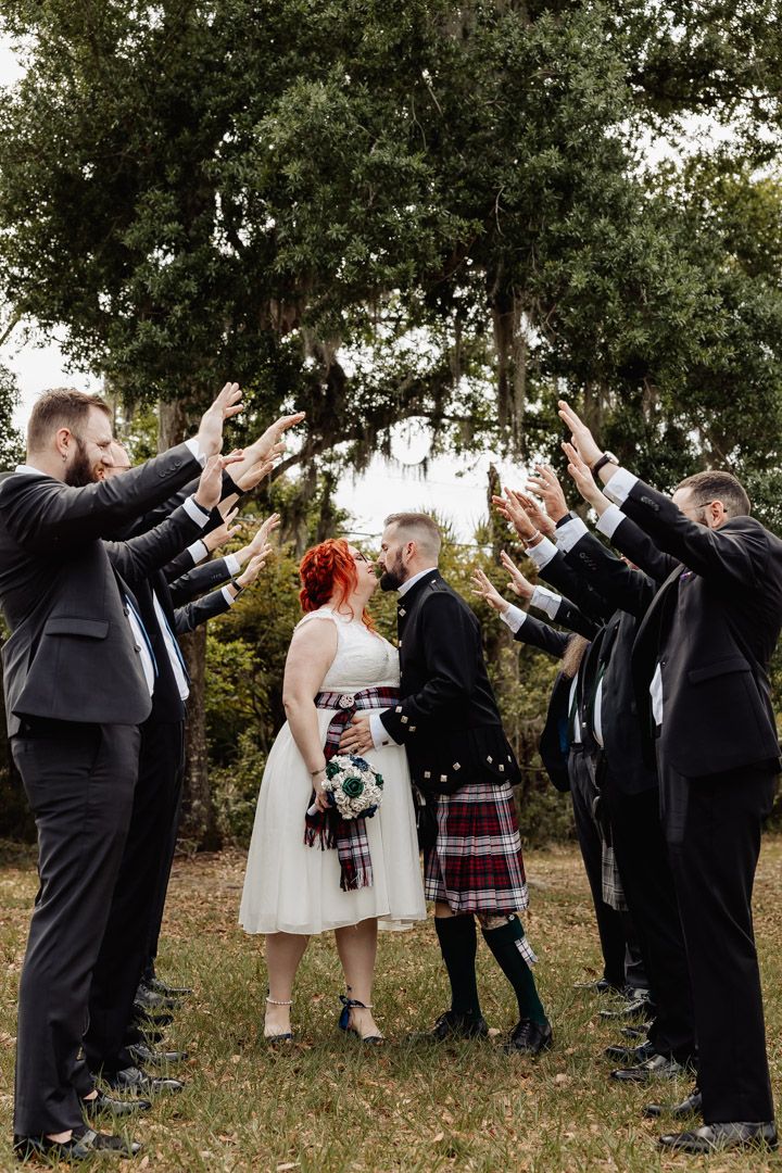 Wedding party forms arch with raised arms as bride and groom share romantic moment beneath canopy of hands.