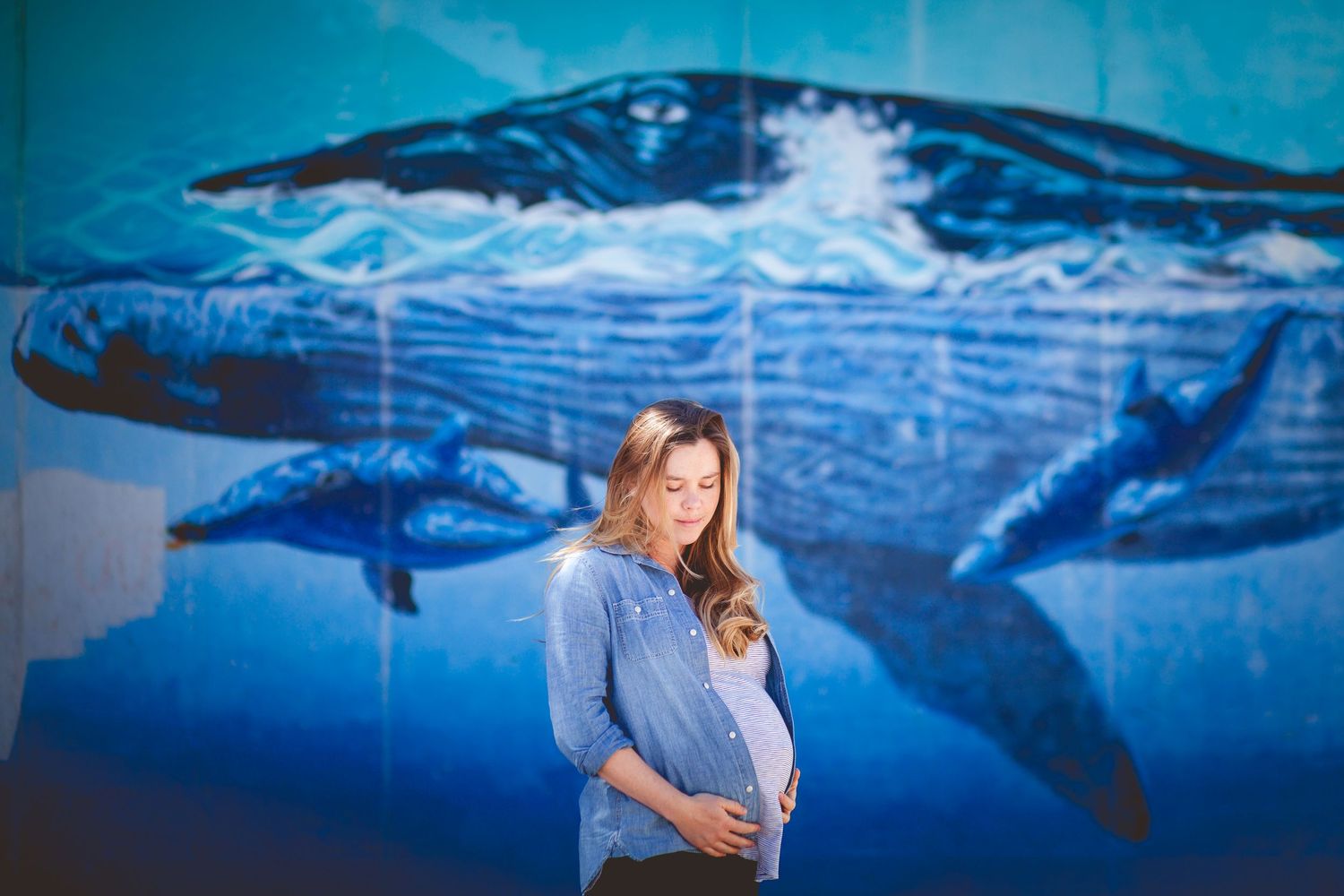 Pregnant woman in blue shirt standing before a vibrant whale mural outdoors.