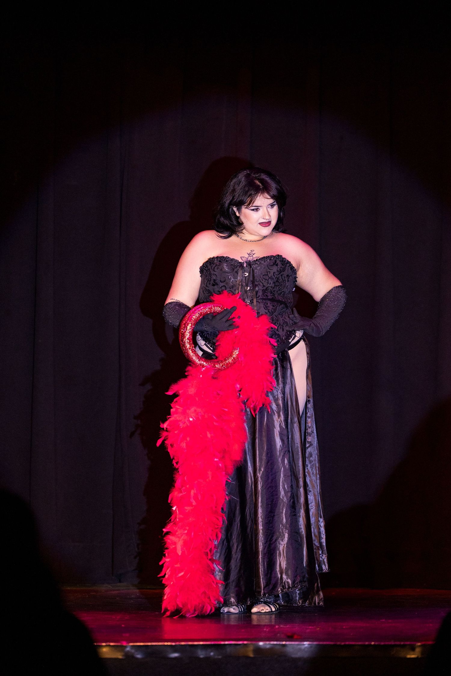 Burlesque performer in black gown with red feather boa on stage under spotlight.