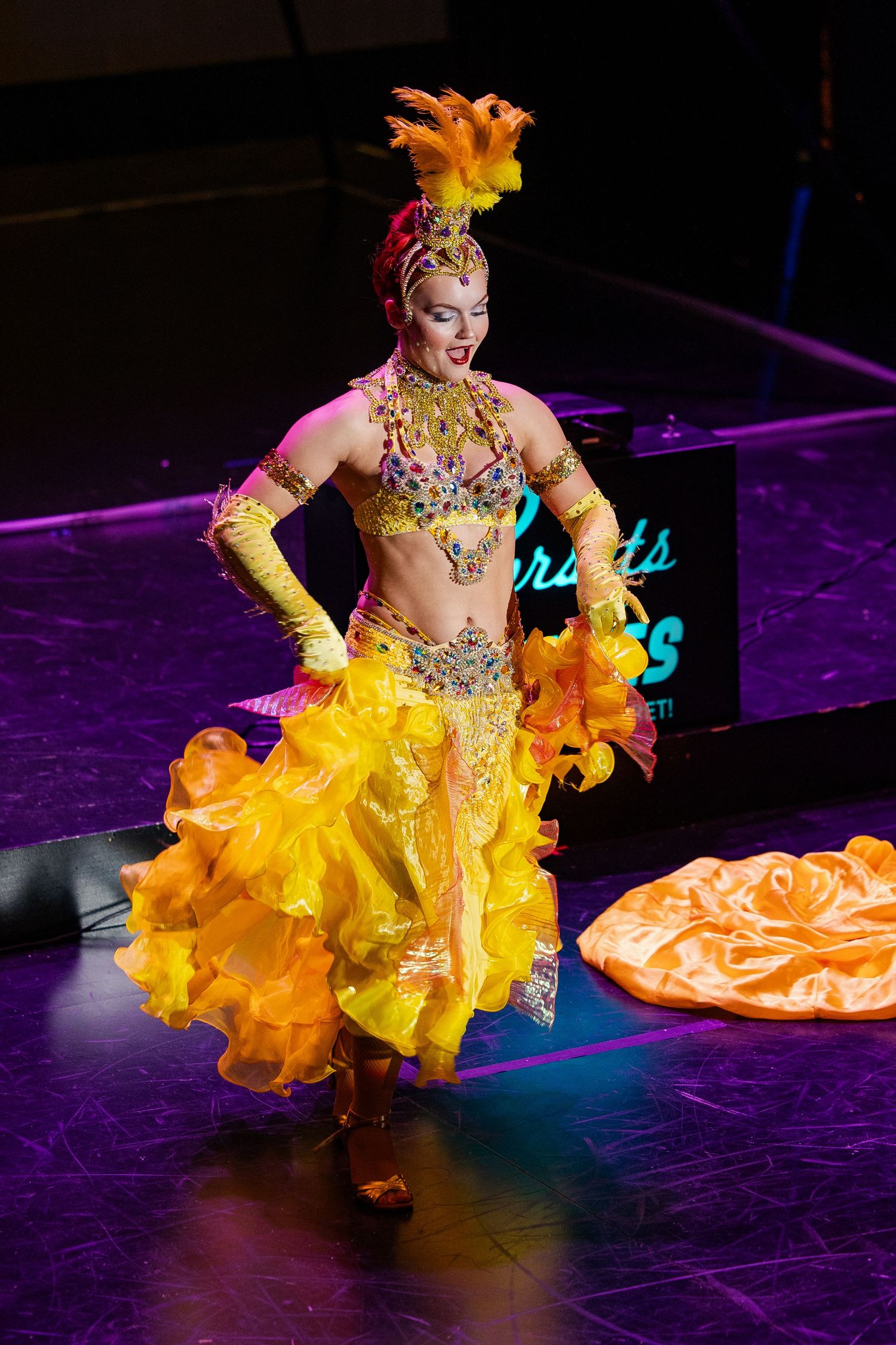 Burlesque performer in ornate yellow and gold costume with feathered headpiece dancing on stage.
