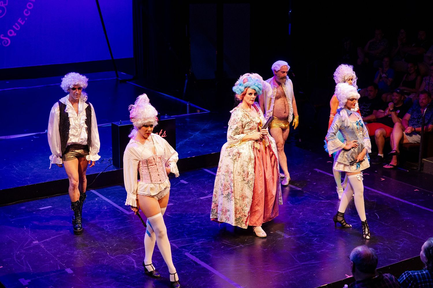 Burlesque ensemble cast in Marie Antoinette-style white wigs and period costumes perform on brightly lit stage.