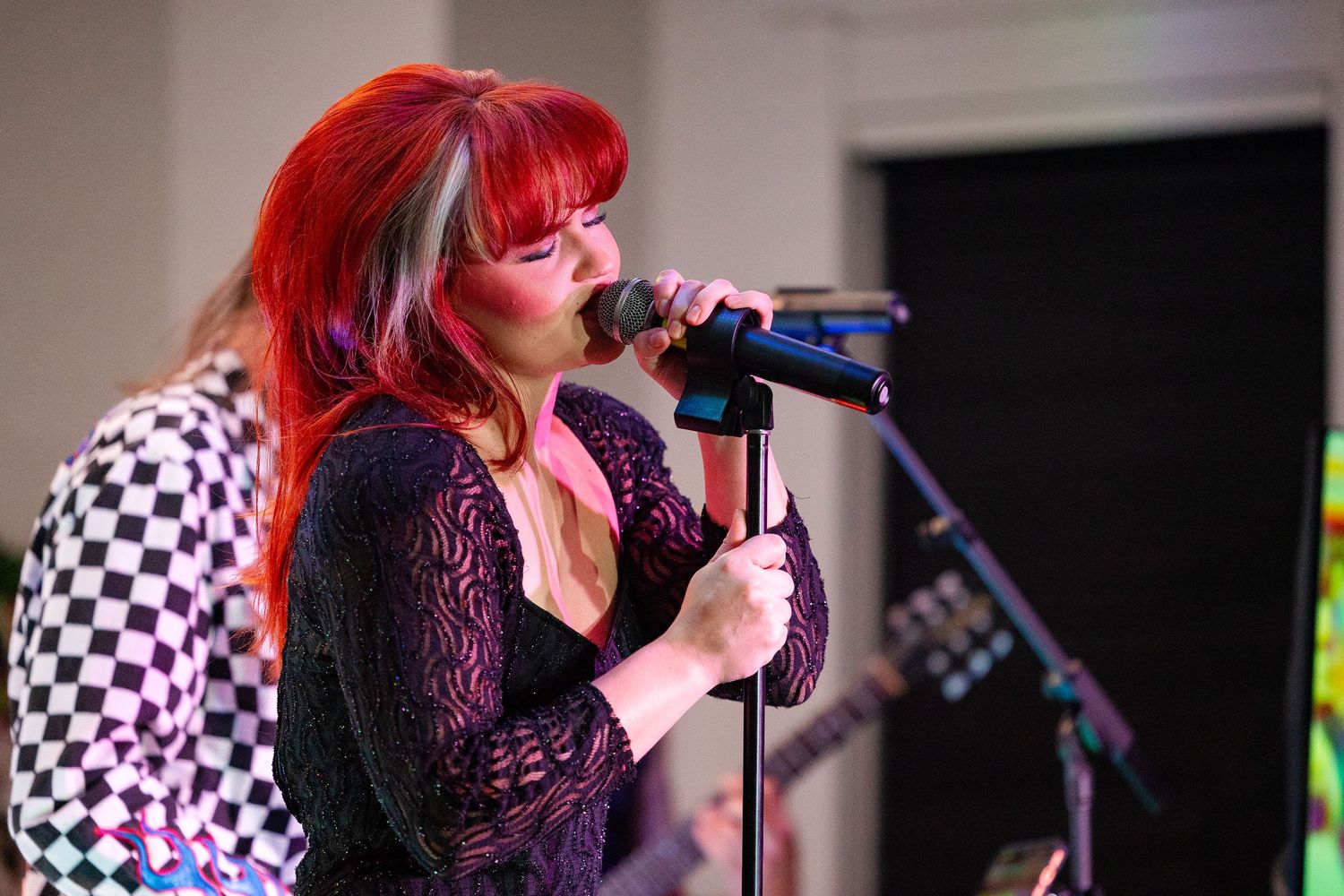 Red-haired female vocalist performing on stage with microphone, wearing black lace top at live music event.