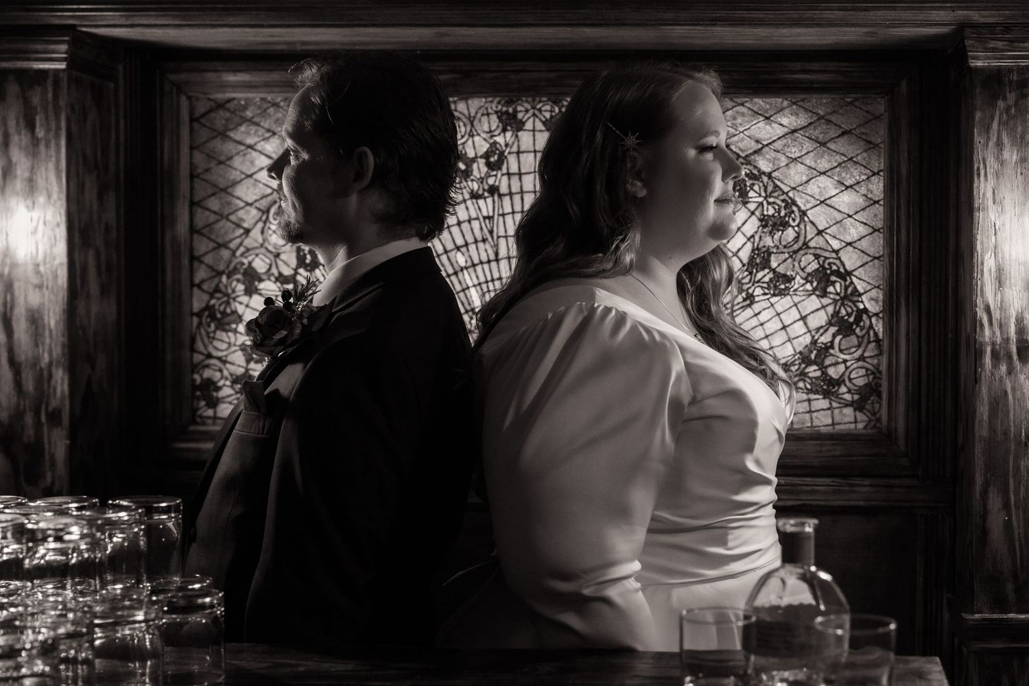 Black and white portrait of bride and groom standing back-to-back at a vintage bar with ornate decor.