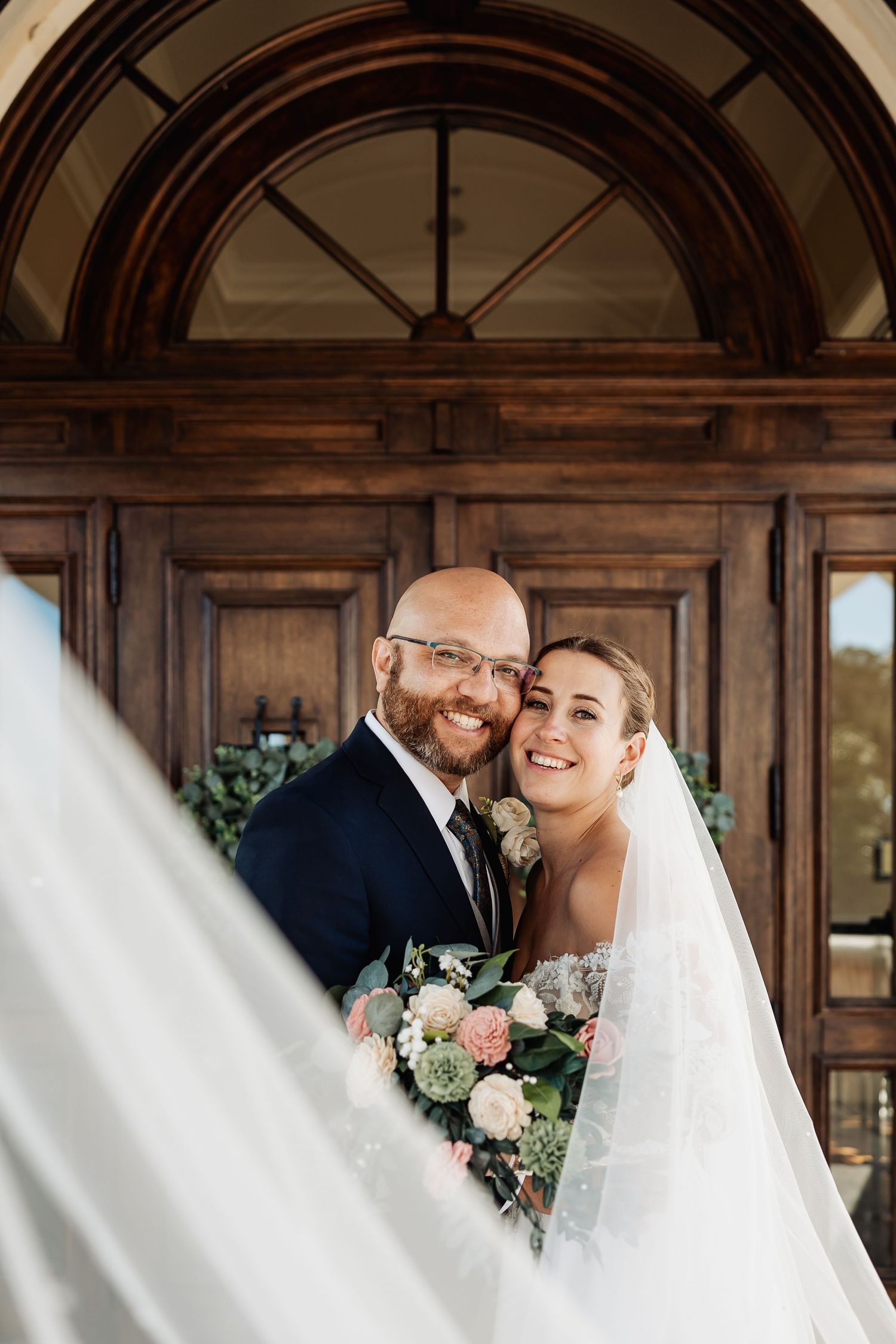 Smiling bride and bald groom embrace before grand wooden doors with flowing veil and bouquet.