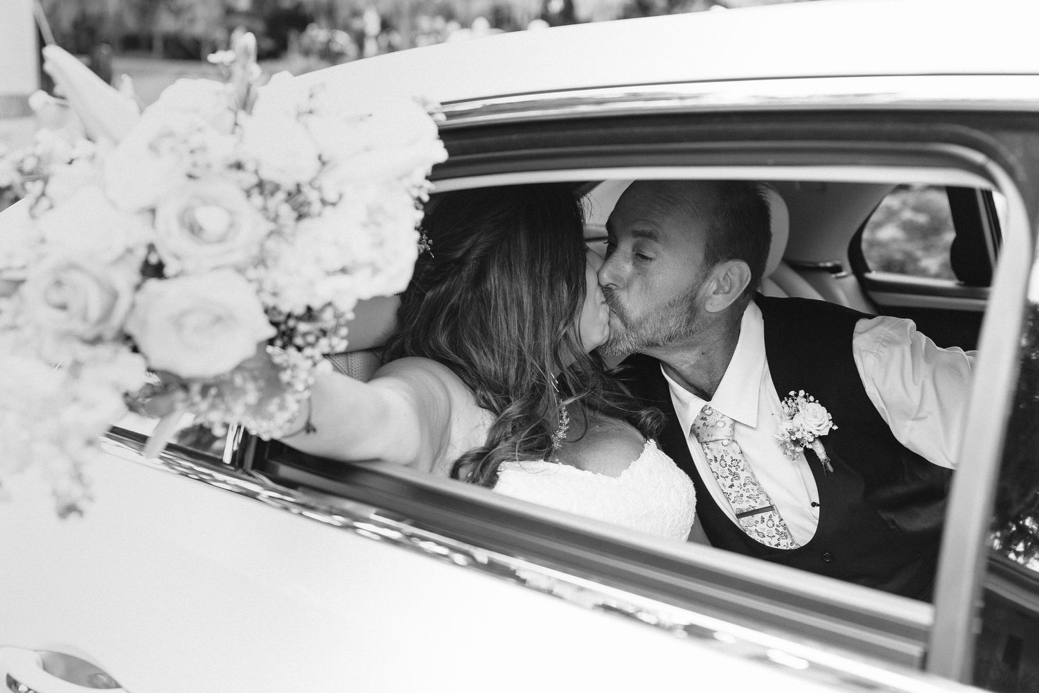 Newlyweds share a passionate kiss inside a classic white car in a romantic black and white wedding photo.