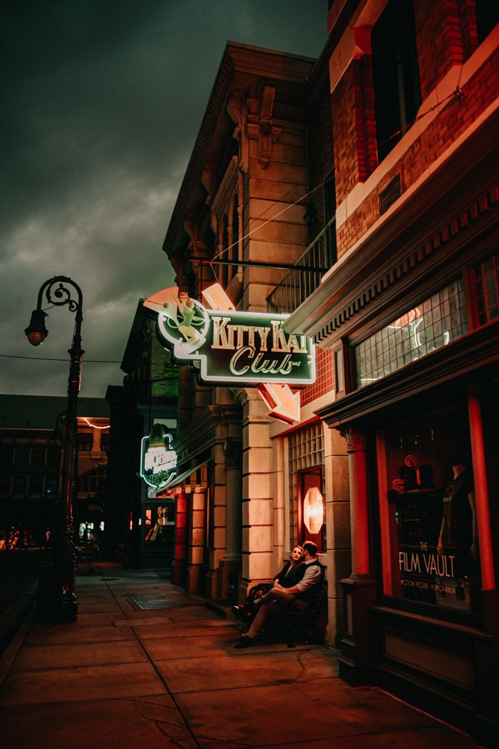 Couple sitting together beneath the Kitty Bar Club neon sign on a moody, dramatic street.