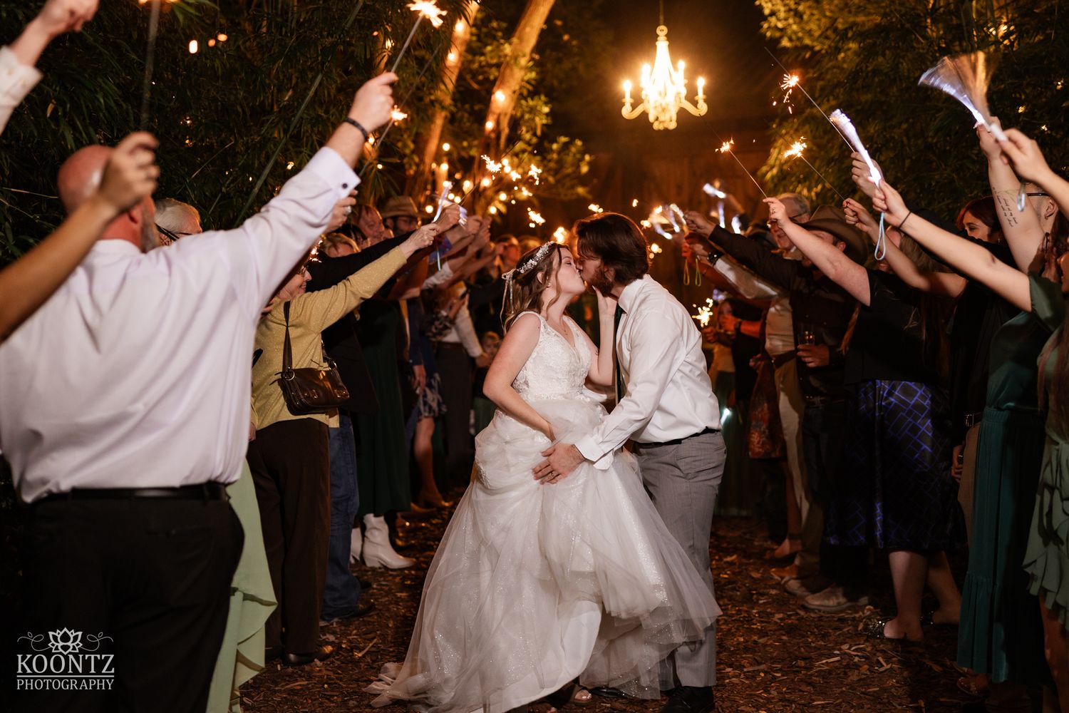Newlyweds share romantic kiss during stunning sparkler sendoff under chandelier at outdoor night wedding.