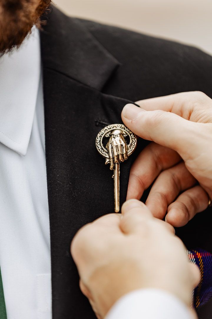Close-up of Hand of the King Game of Thrones brooch being pinned to groom's black jacket.