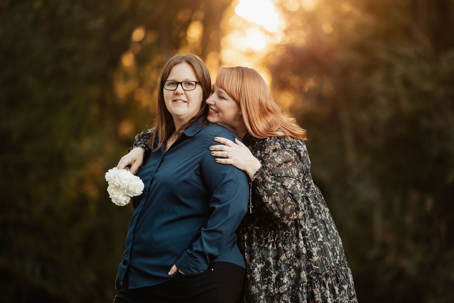 A woman in a floral dress kisses her partner on the cheek while hugging her from behind in a golden forest.
