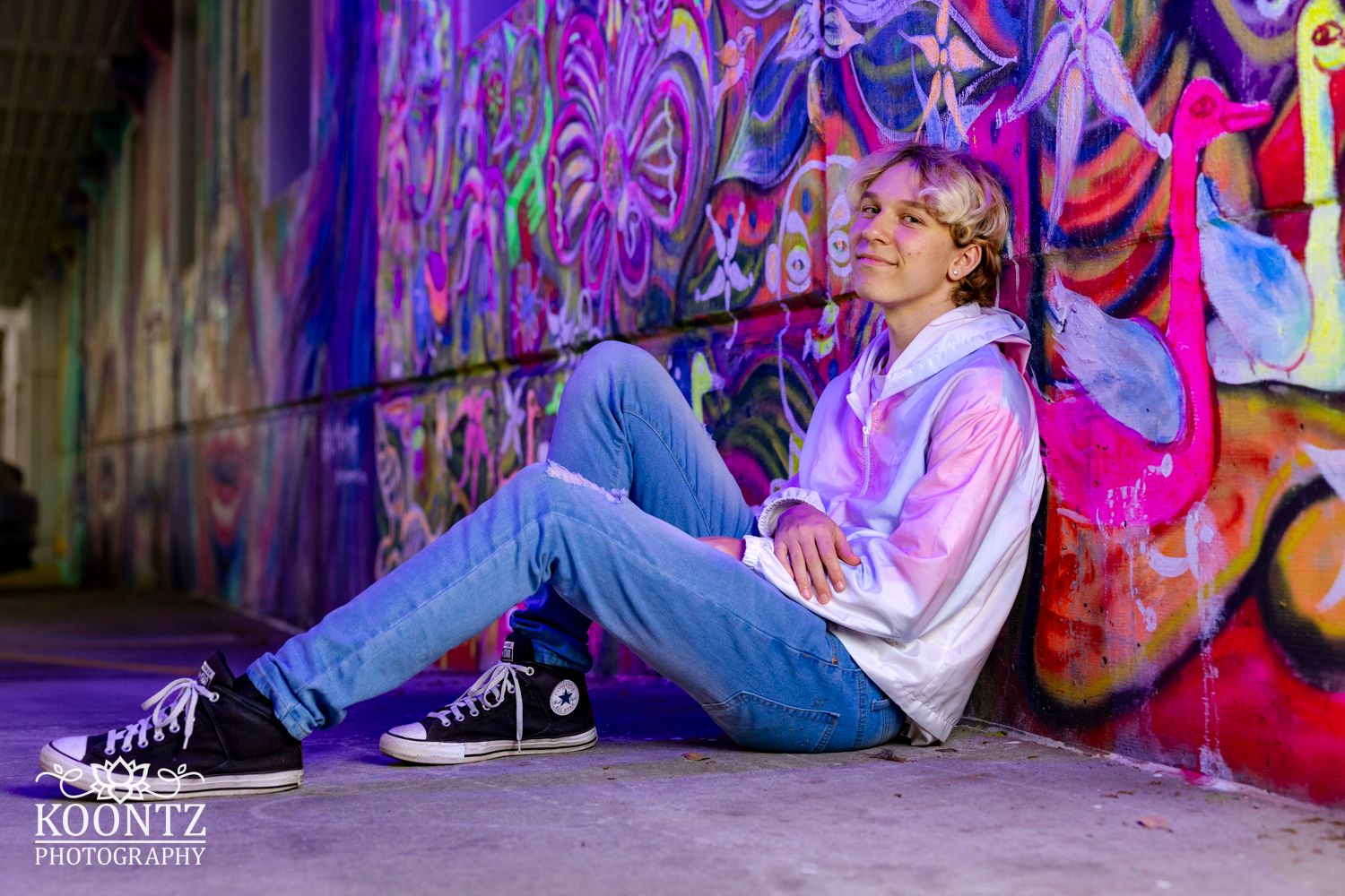 Person in white hoodie and jeans sits casually against colorful street art in purple lighting.