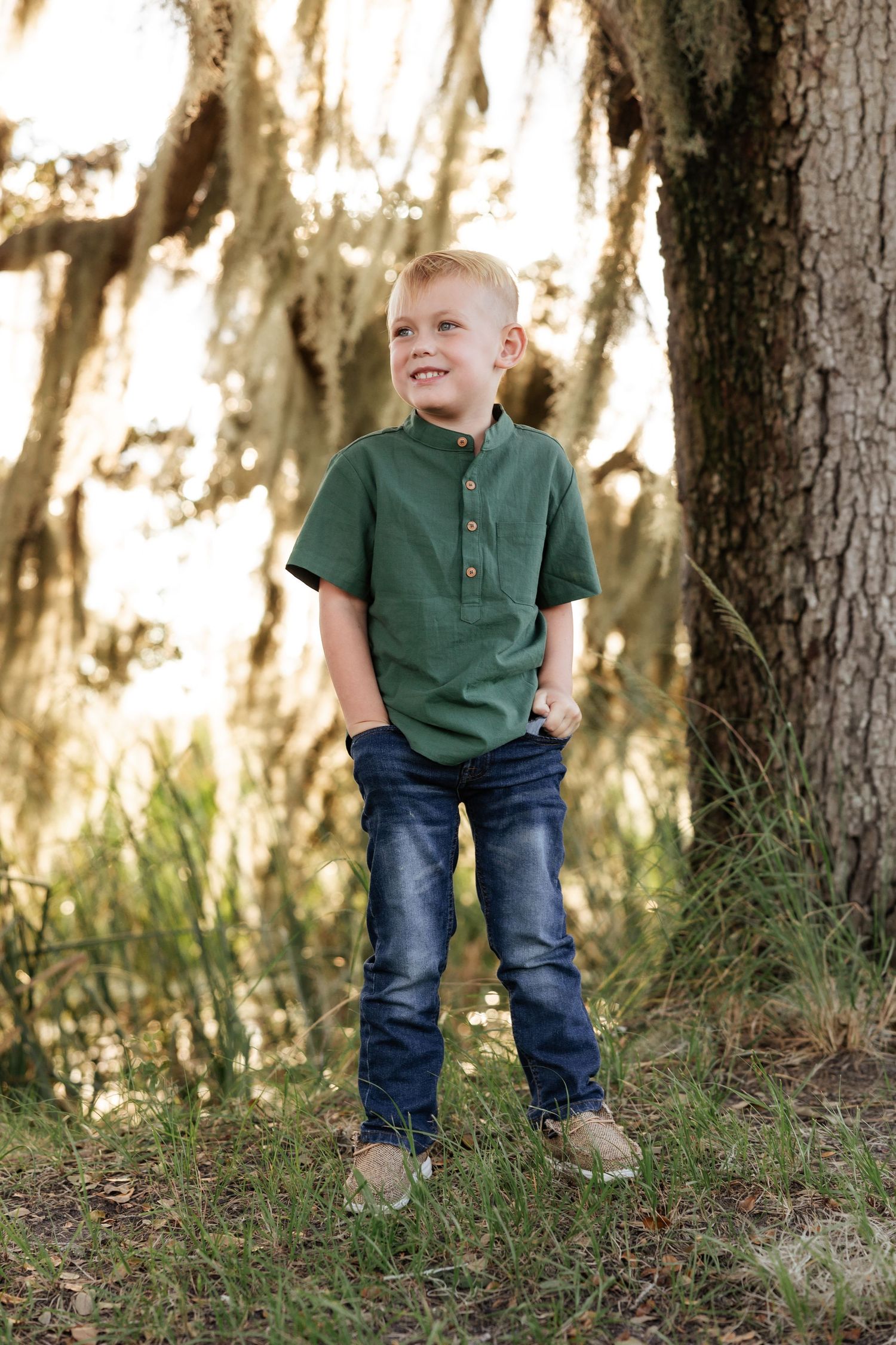 Child in green shirt and jeans stands confidently by large tree trunk.