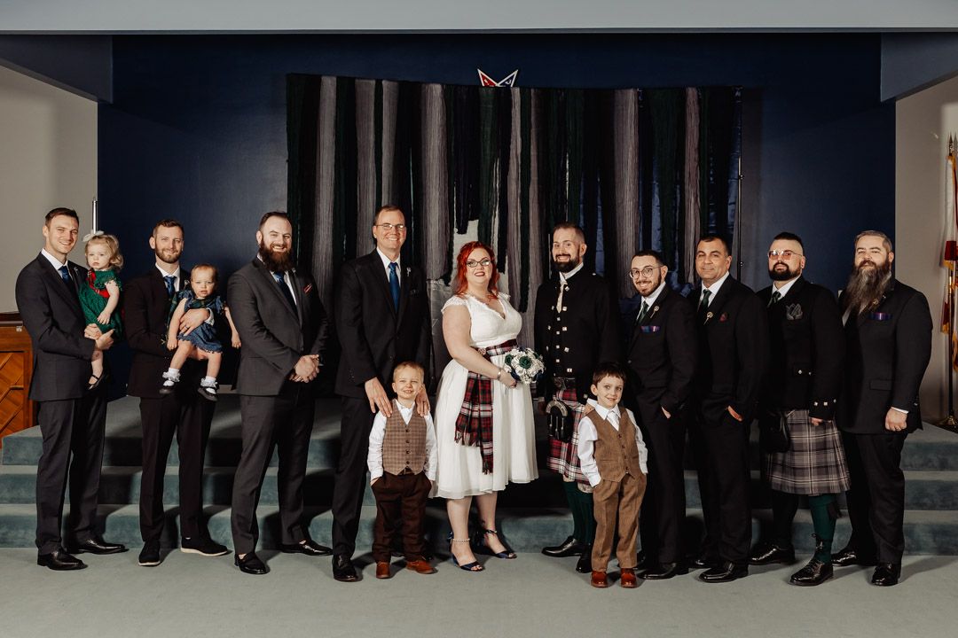 A wedding party group photo featuring formal attire and traditional Scottish kilts indoors.