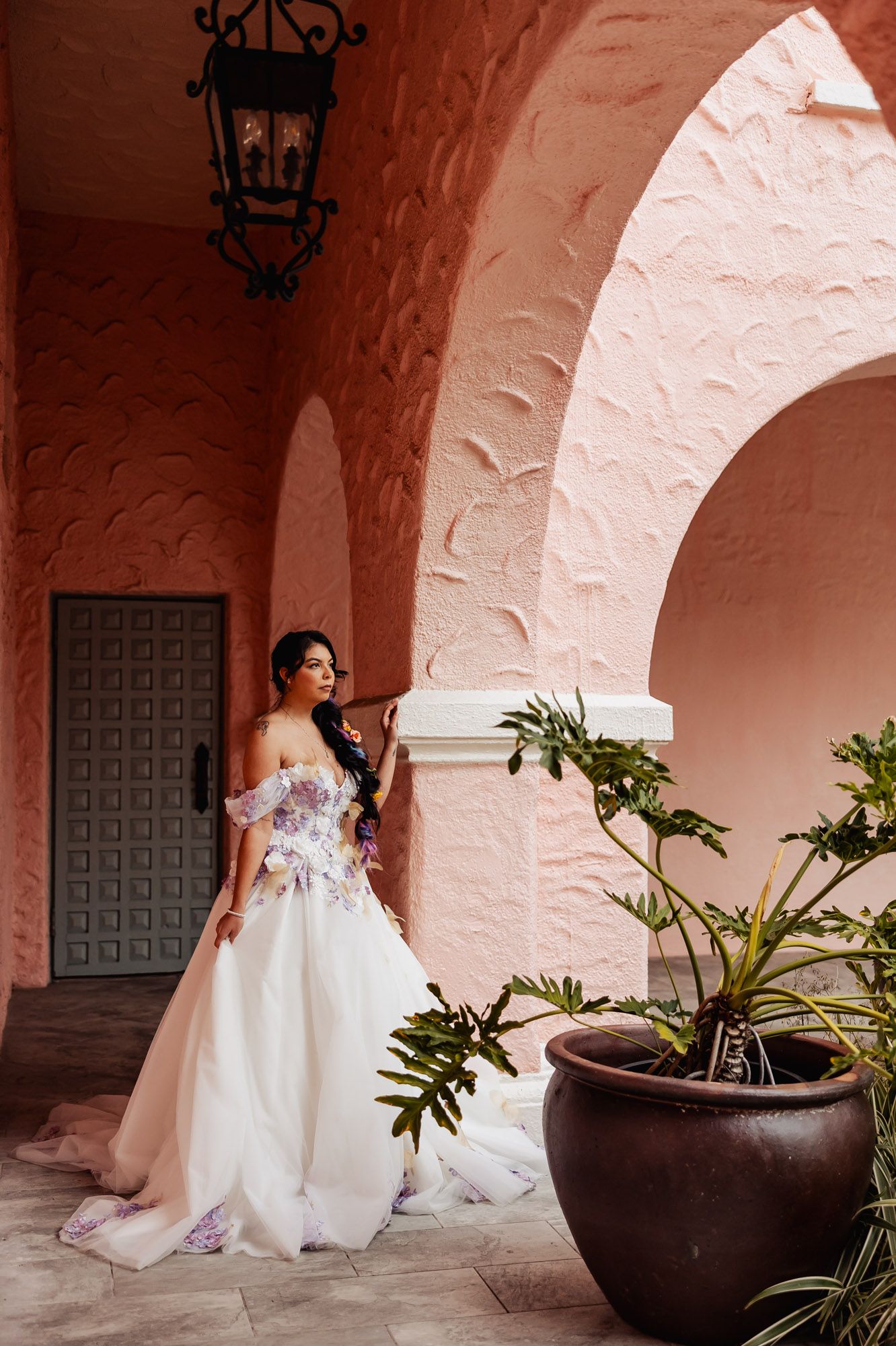 Bride in white floral off-shoulder gown poses under pink stucco archway beside large decorative planter.