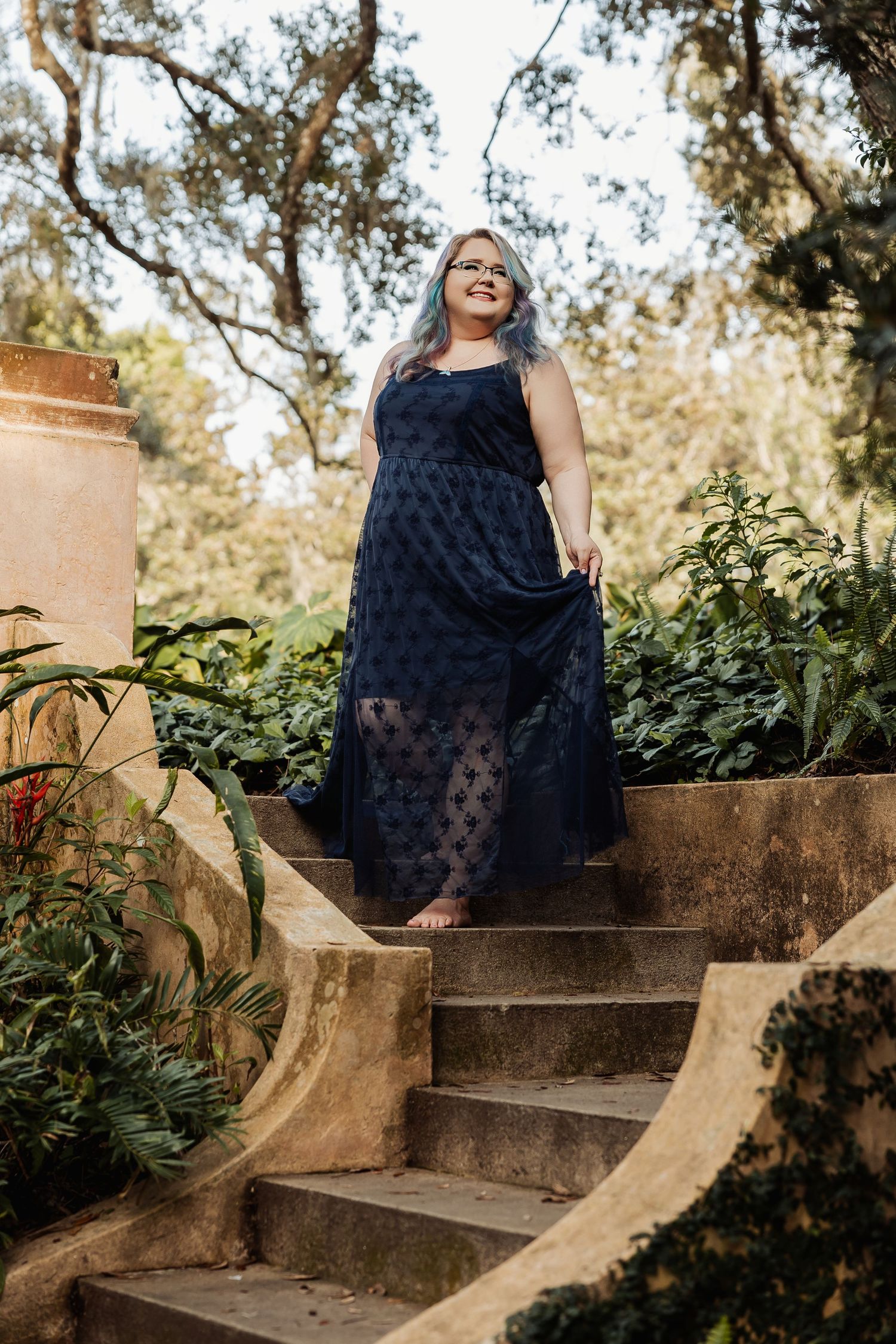 Woman in navy lace gown stands on stone garden steps surrounded by lush greenery.