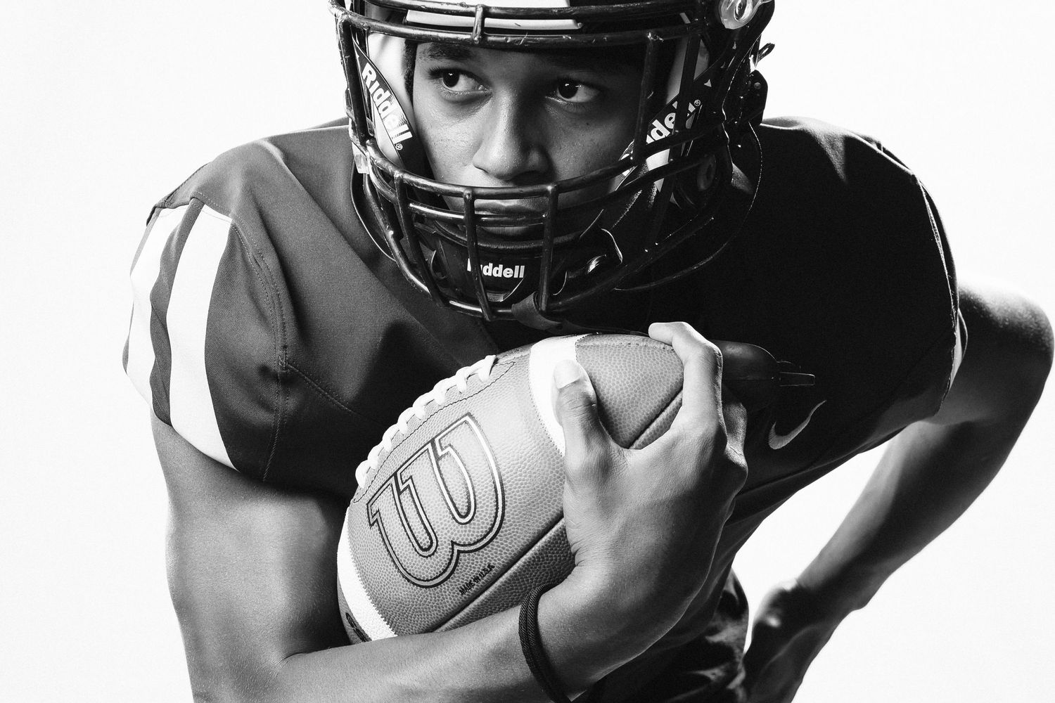 Black and white photo of a young football player in helmet holding a Wilson football in a defensive pose.