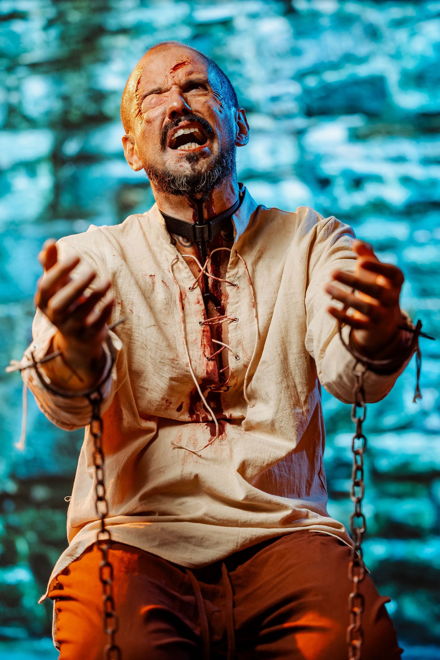 Anguished man in bloody costume with chains raises hands in dramatic theatrical or Halloween performance.