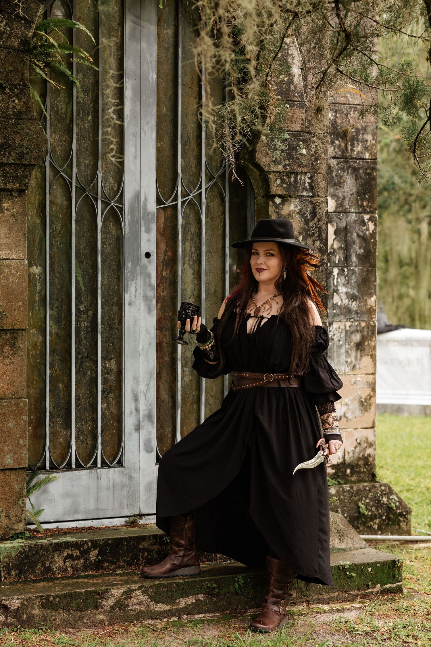Woman in black witch dress and hat leans against mossy stone gate holding binoculars outdoors.