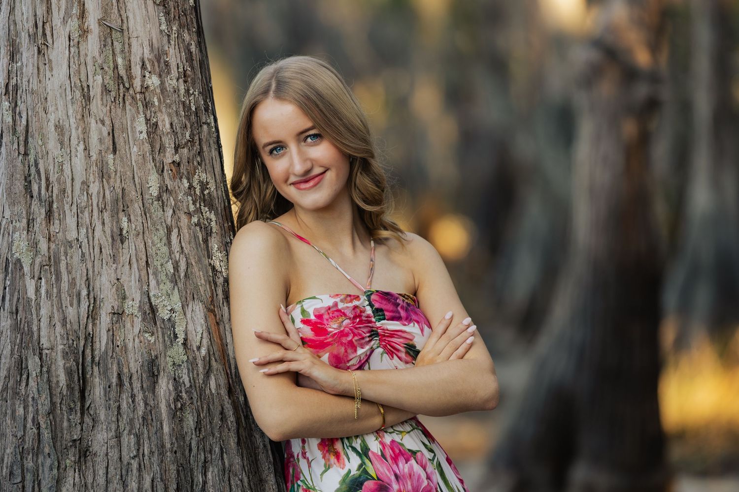 Young woman in floral dress leaning against tree trunk smiling outdoors.
