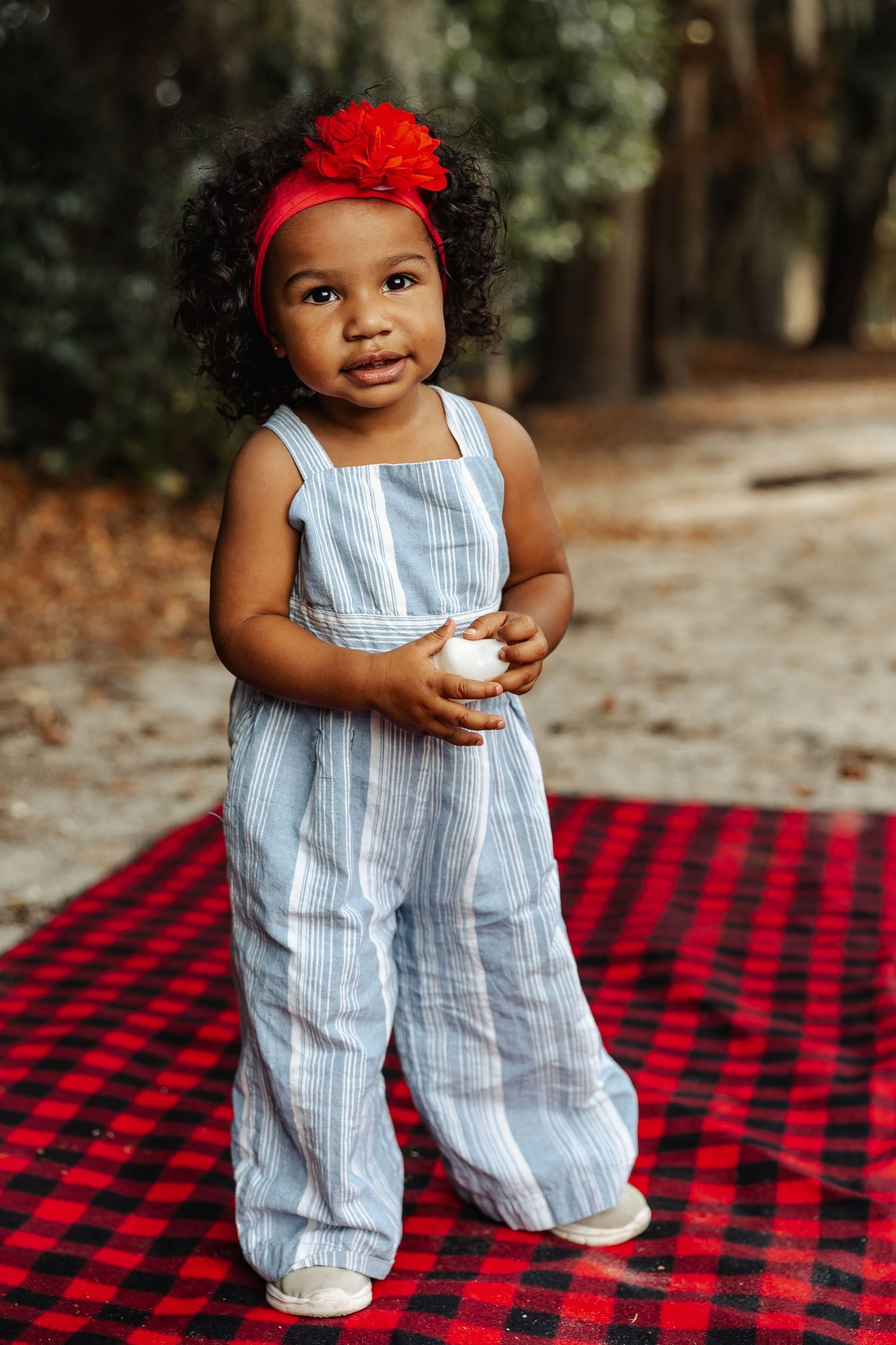 A cute toddler girl in a striped jumpsuit with a red bow stands on a plaid blanket.