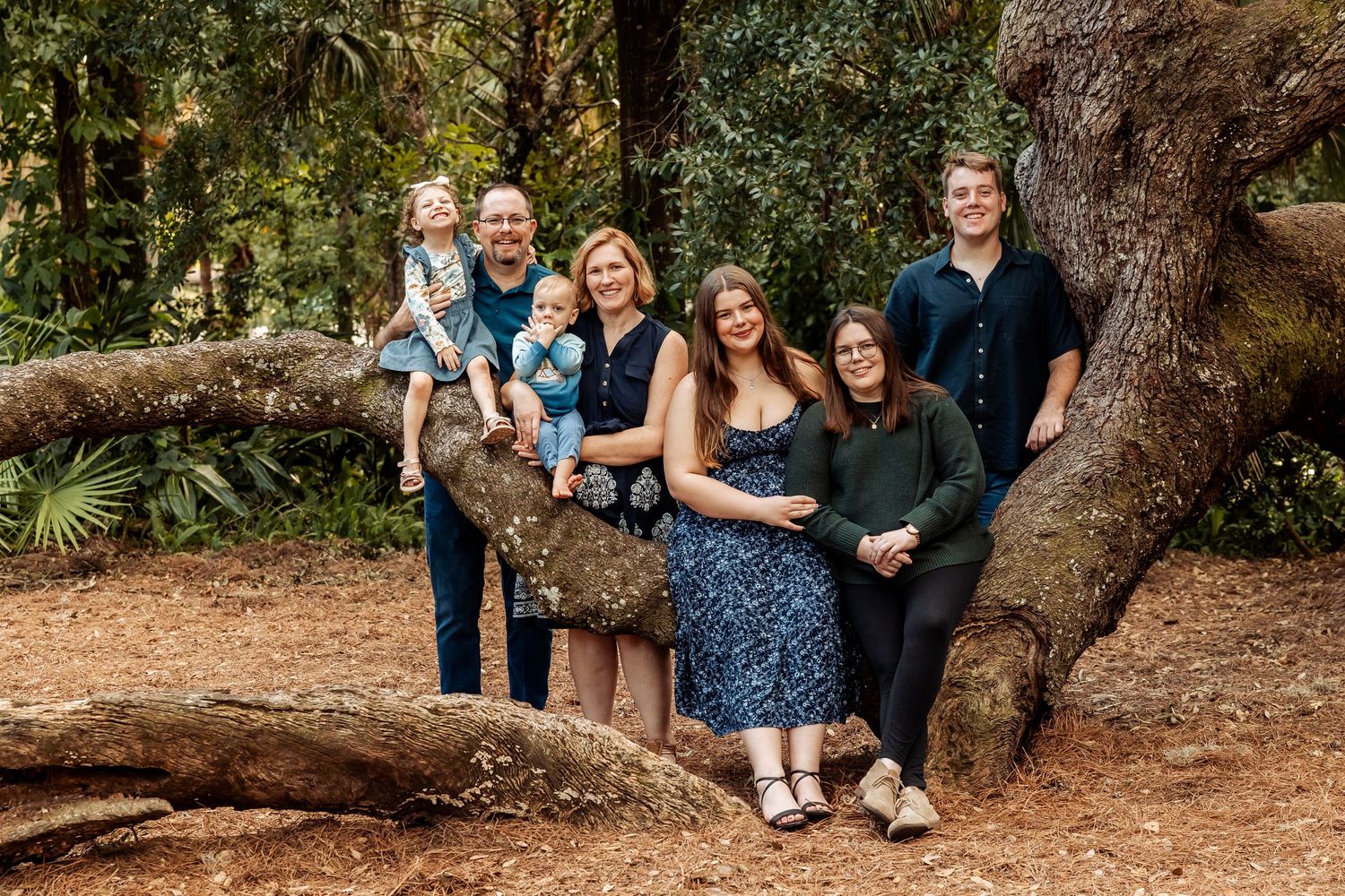 Extended family of seven posing together near a large tree in a sunny outdoor park setting.