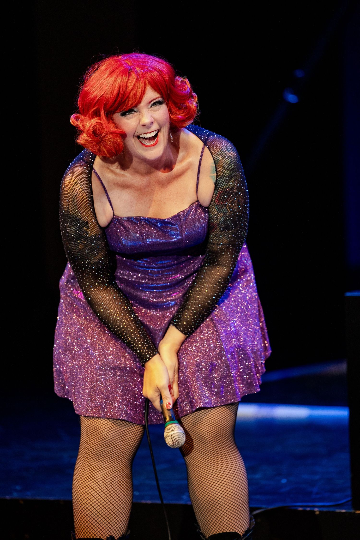 Energetic red-haired performer in purple sequin dress laughing on stage during burlesque show.