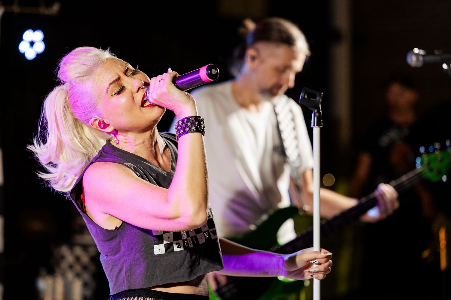 Blonde female singer performs passionately under pink stage lights with guitarist behind her.