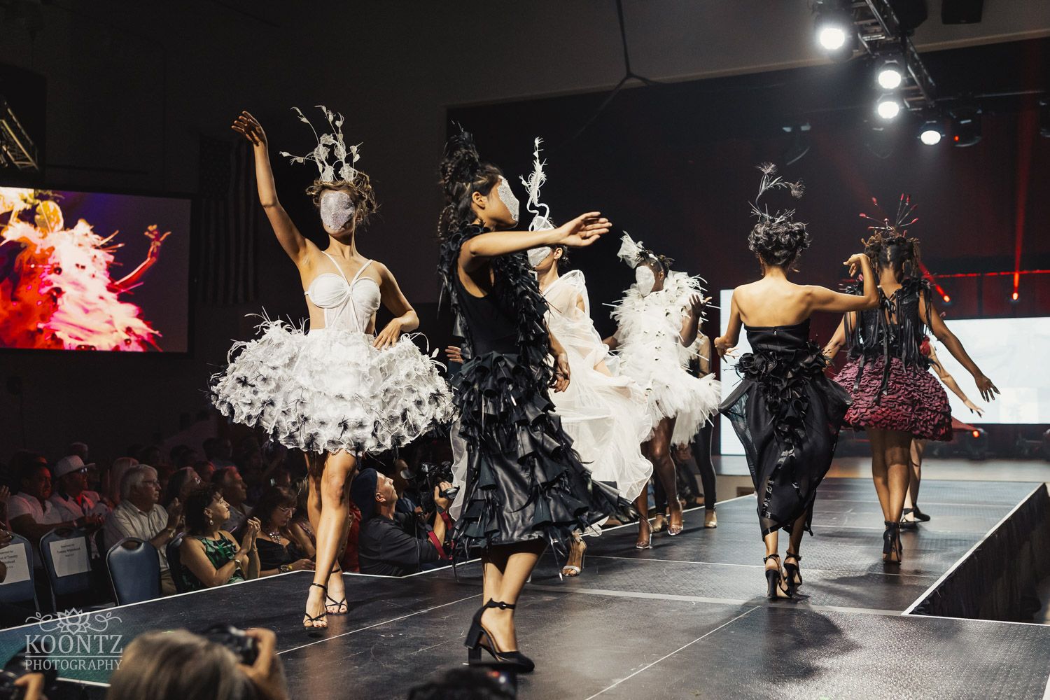 Fashion show runway featuring models walking in formal attire under dramatic lighting and beneath neon displays.