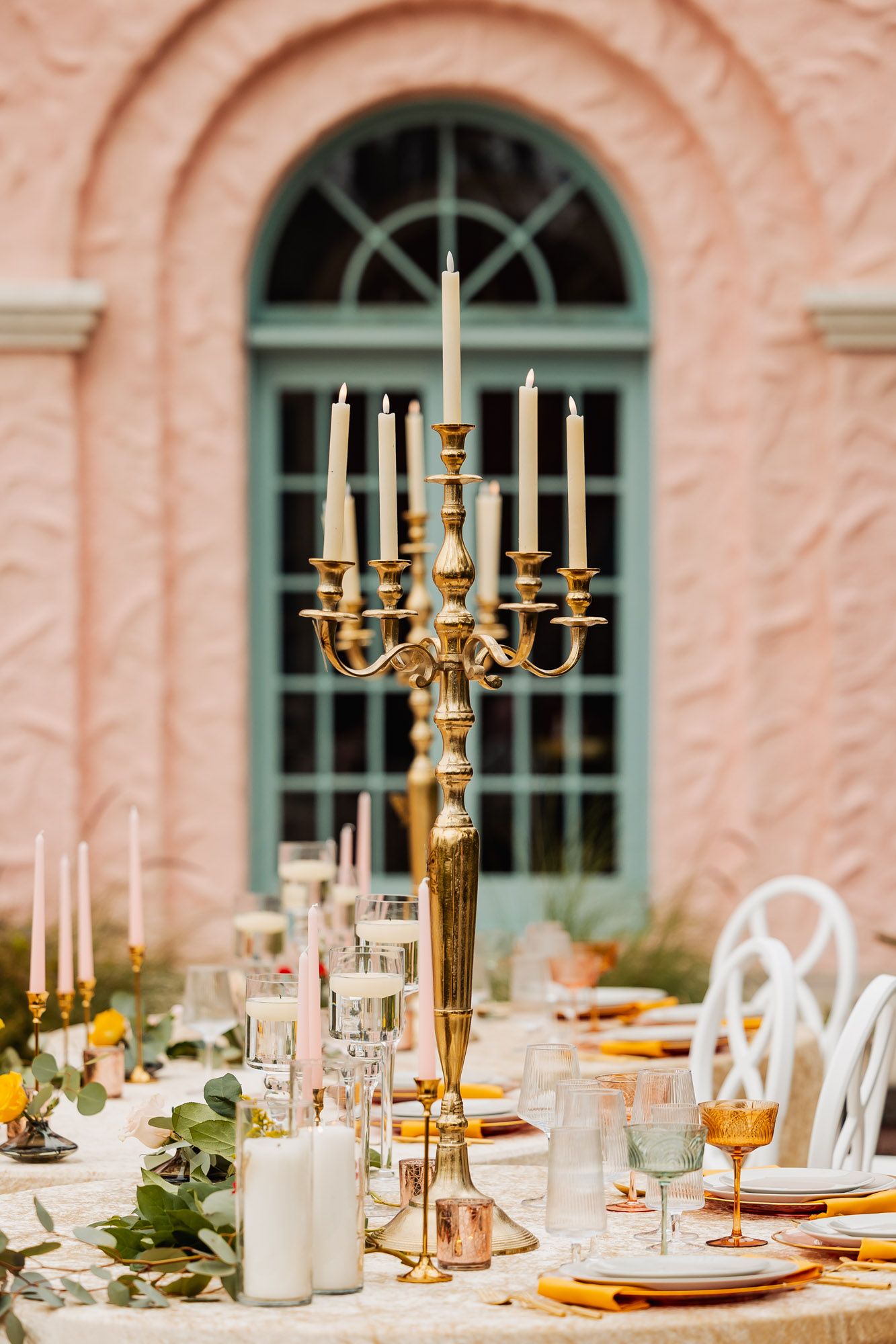 Elegant wedding table setting with gold candelabra centerpiece and white candles against a pink building with arched window.