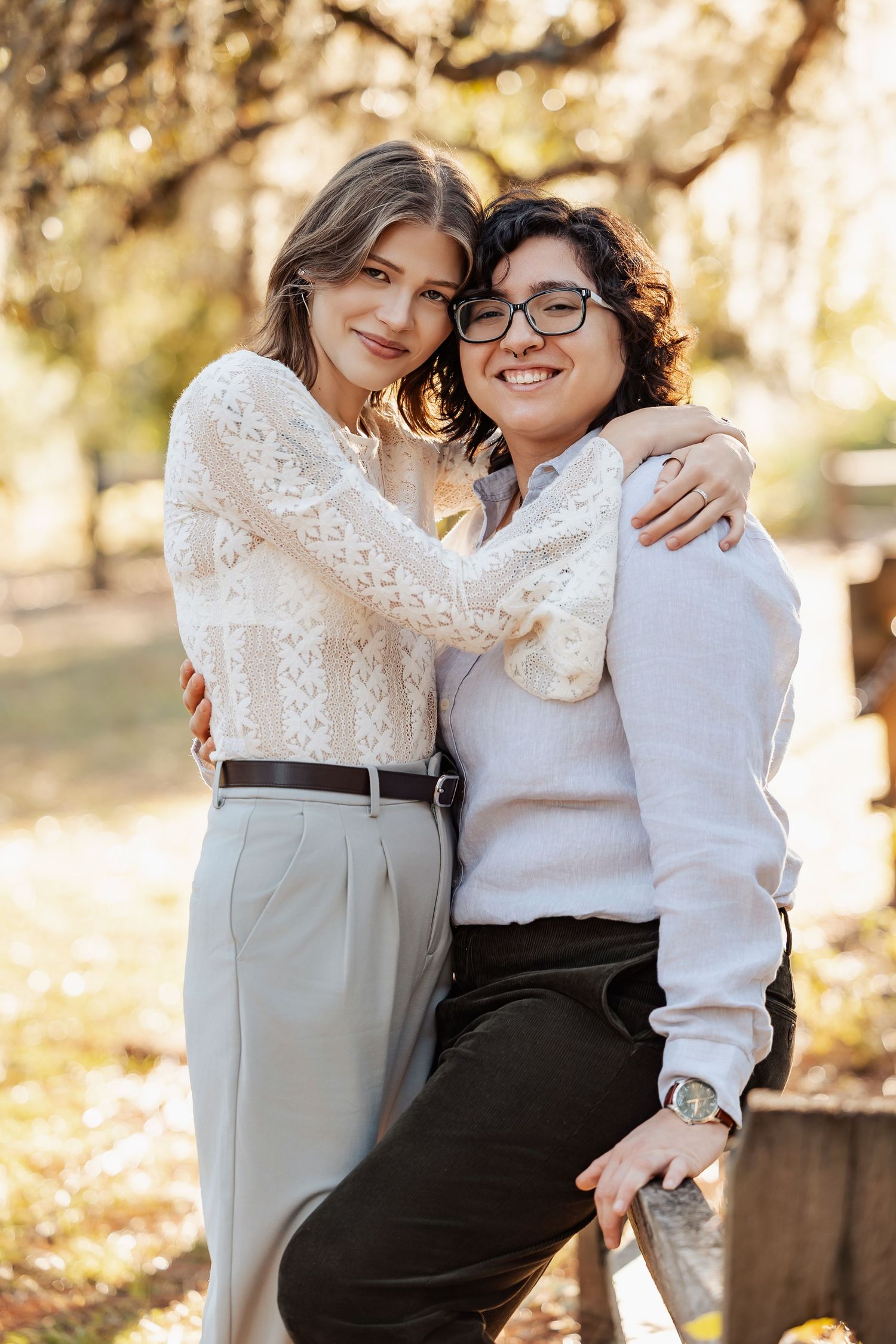 Two women hugging and smiling on a wooden fence in a sun-dappled outdoor park setting.
