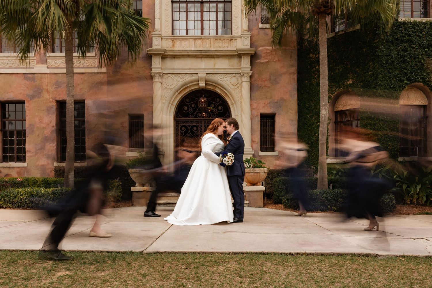 Bride and groom share a romantic kiss while wedding party rushes past in a motion-blur photo outside a historic venue.