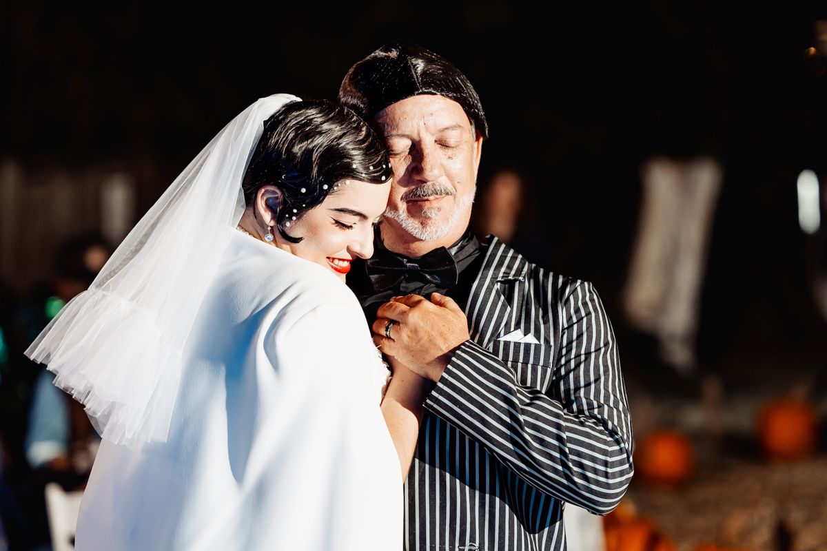 Bride and older man sharing a warm embrace at an outdoor Halloween-themed wedding reception.