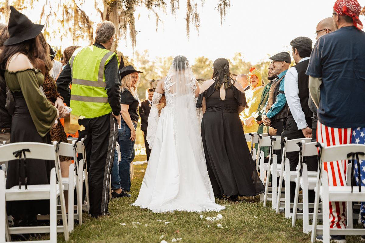 Bride walking down the aisle in white gown and veil at outdoor Halloween-themed wedding ceremony.