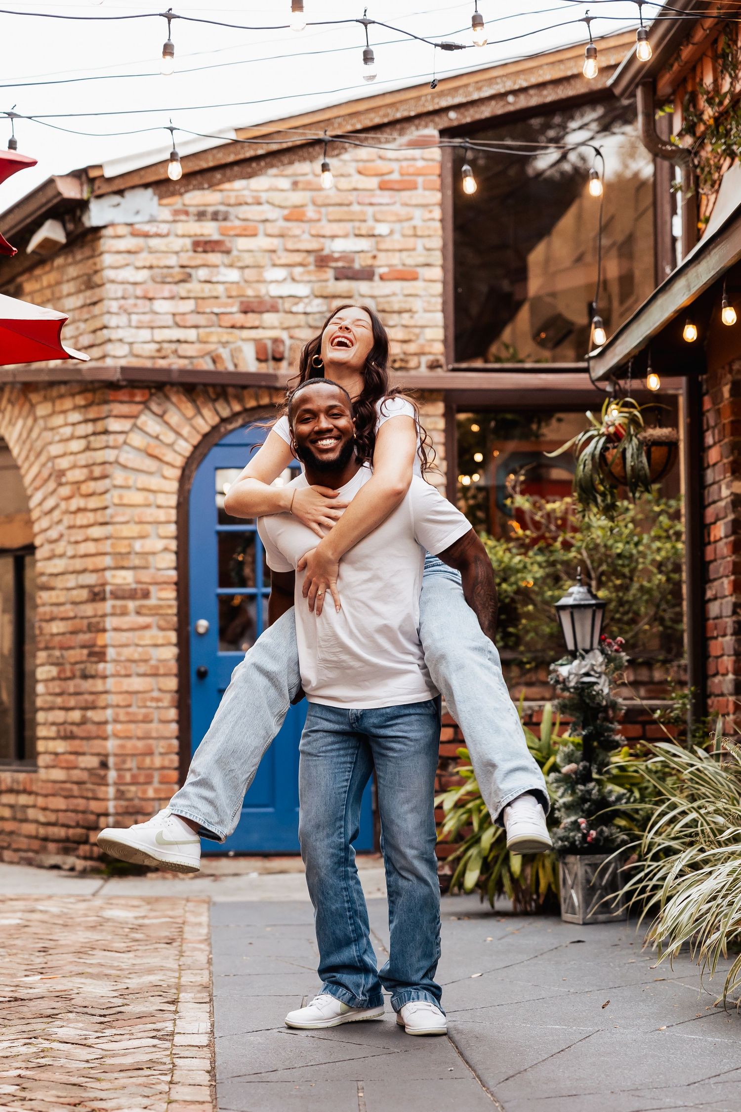 Smiling man giving laughing woman a piggyback ride outside a charming brick building.