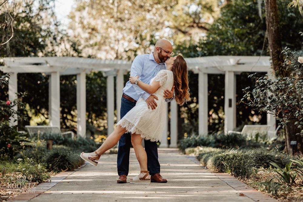 A romantic dip pose captured in front of a white pergola in a manicured garden setting.